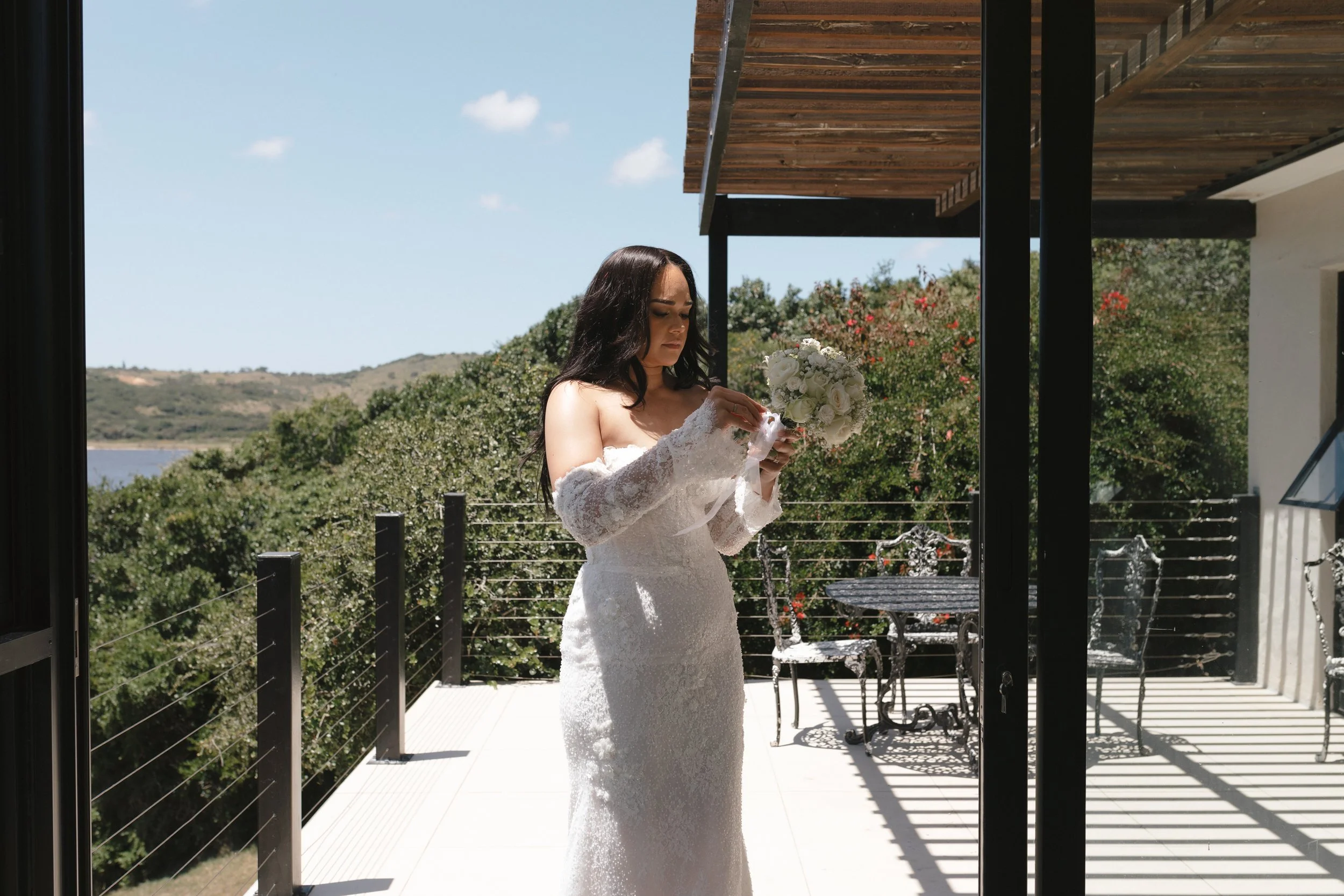 A woman in a wedding dress holds a bouquet of white flowers on a balcony with a scenic outdoor view, under a wooden pergola, during daytime. Gqeberha wedding photographer at Royalston Lakehouse.