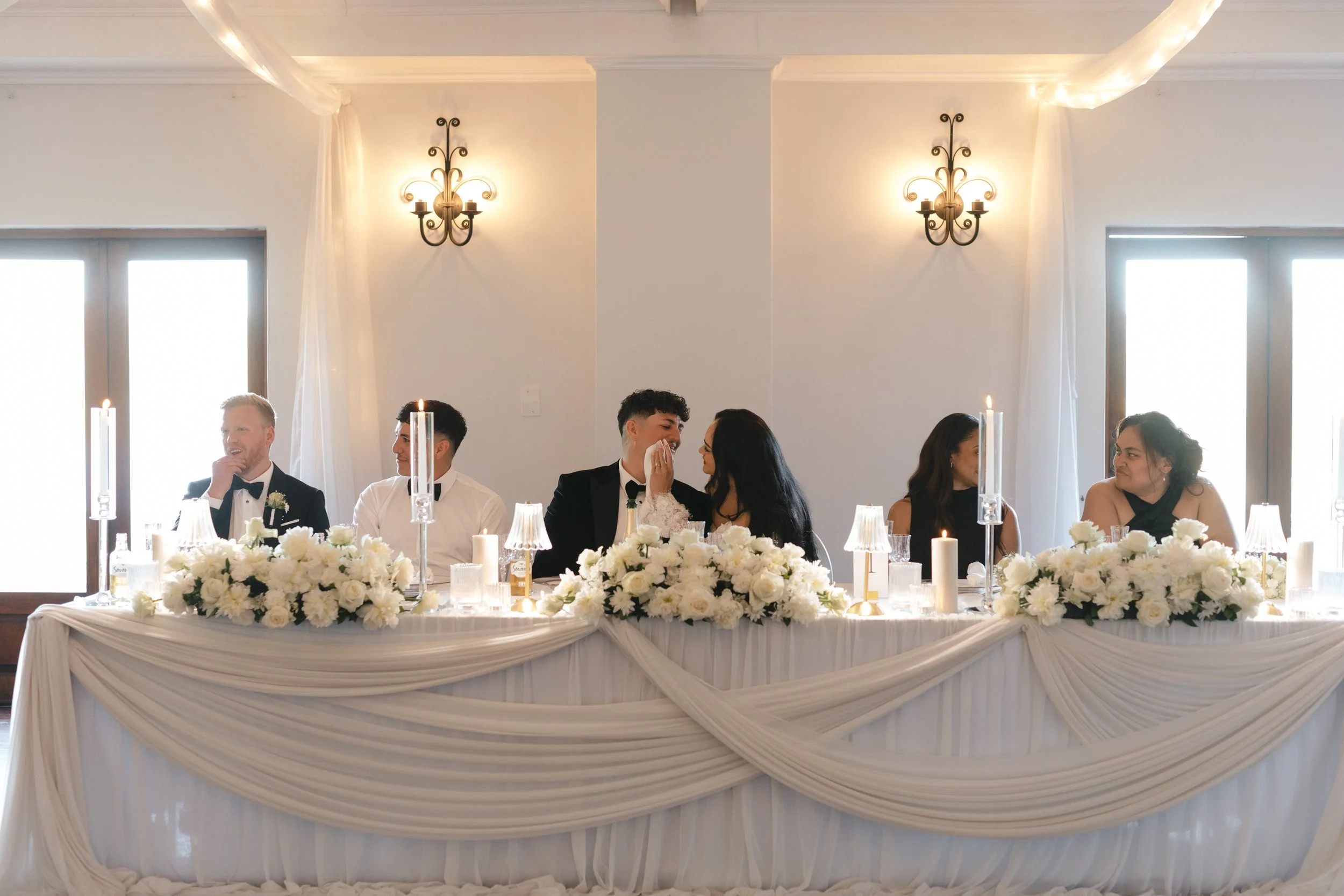 A wedding reception table decorated with white flowers, candles, and draped white fabric. Guests are seated at the table, engaged in conversation and smiling. Gqeberha wedding photographer The Hire Warehouse - Floral Creations