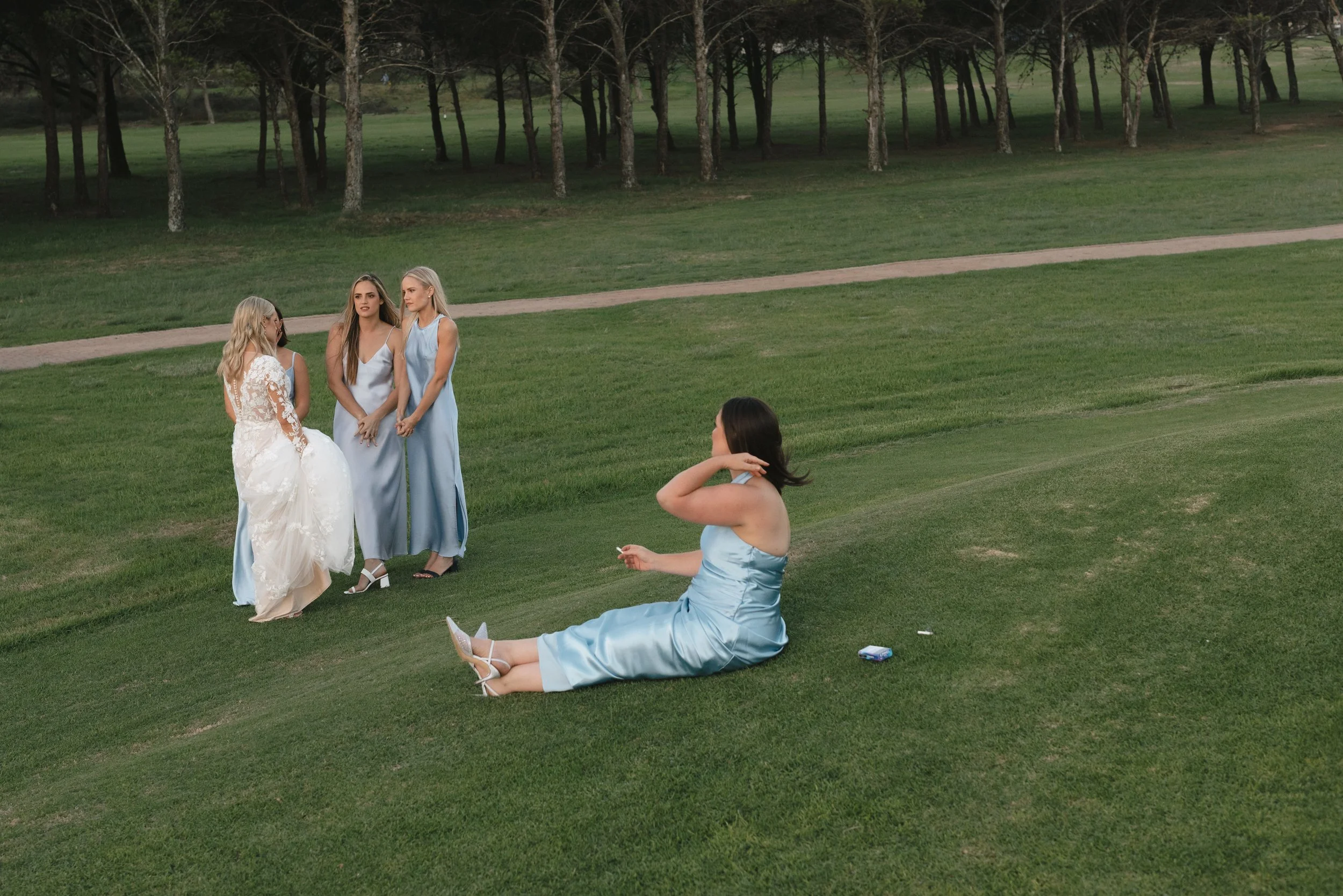 Four women in pastel dresses, one in a wedding dress, standing and sitting on a grassy area in a park with trees in the background, possibly during a wedding or photoshoot. Port Elizabeth Golf Club, Gqeberha