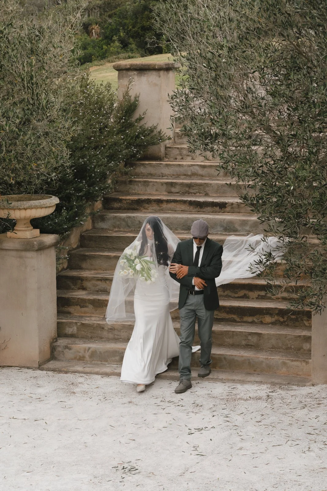 A bride in a white wedding dress with a veil holding a bouquet of white flowers, walking with a man in a black blazer, gray pants, and a gray hat down stone stairs outdoors, surrounded by greenery. Gqeberha wedding photography at The Rose Barn