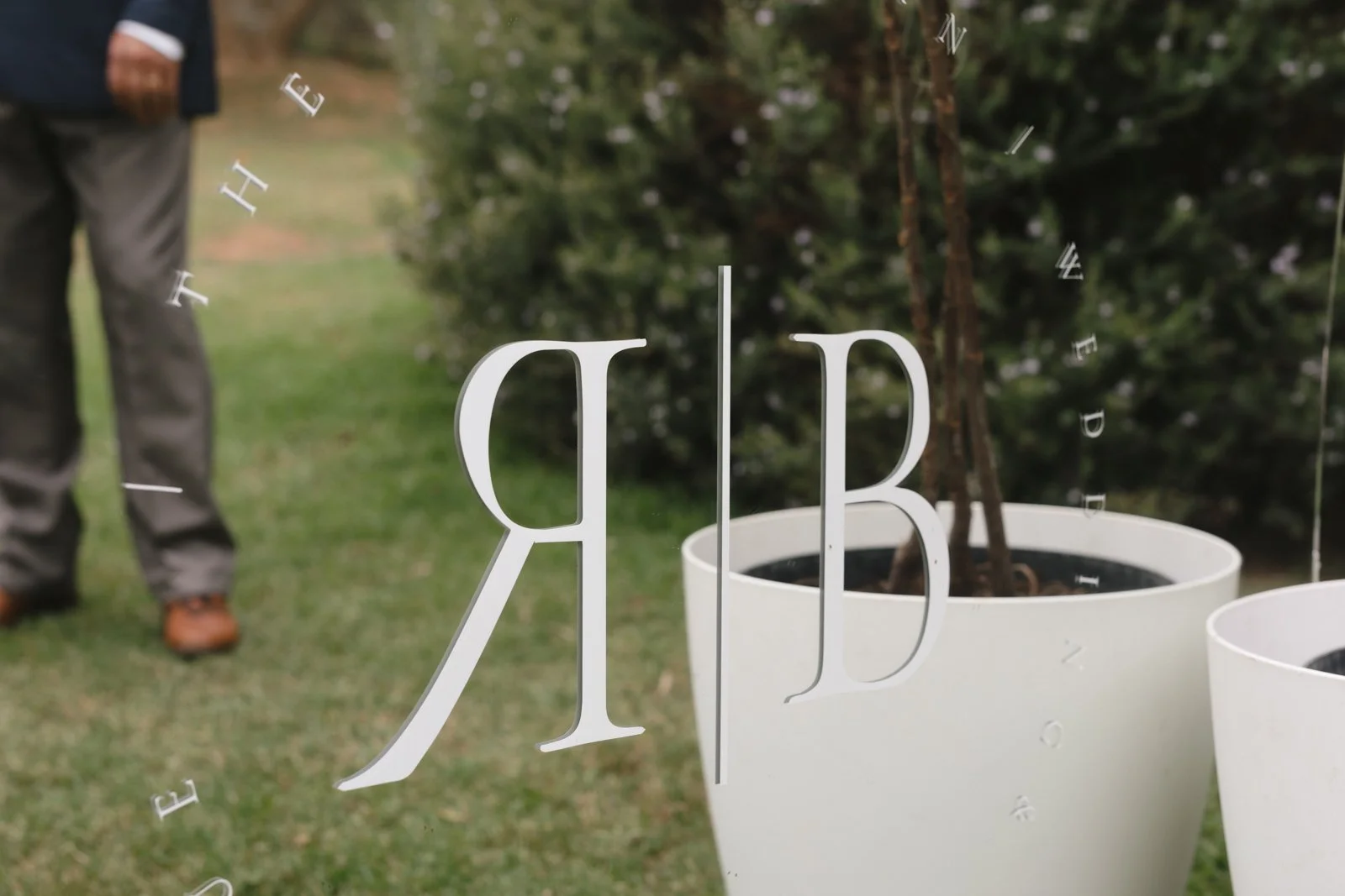 Close-up of a mirror with decorative letters, reflecting a person standing outdoors next to potted plants. Gqeberha wedding photography at The Rose Barn, Jefferys Bay, dress by Werner Dey