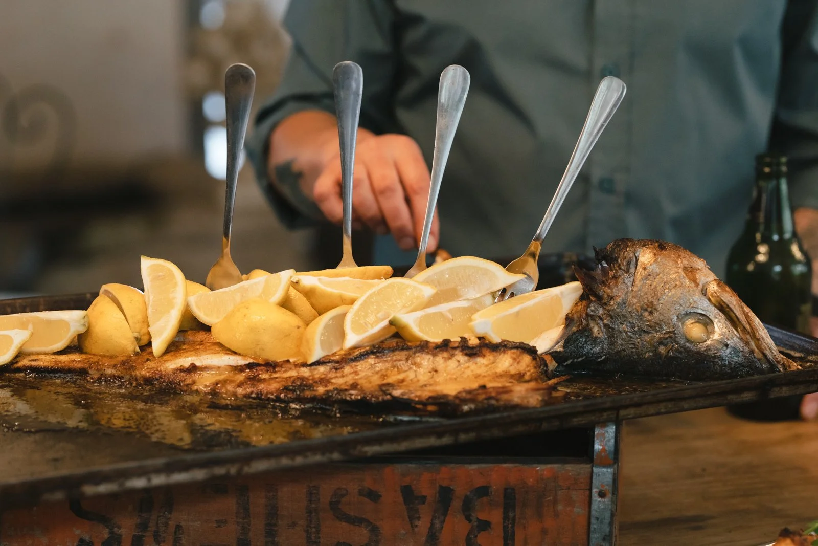 Grilled fish topped with lemon wedges on a tray with a person's hand in the background. Gqeberha wedding photography at The Rose Barn, Jefferys Bay, Lelik Maar Lekker (Chef Gert Joubert)