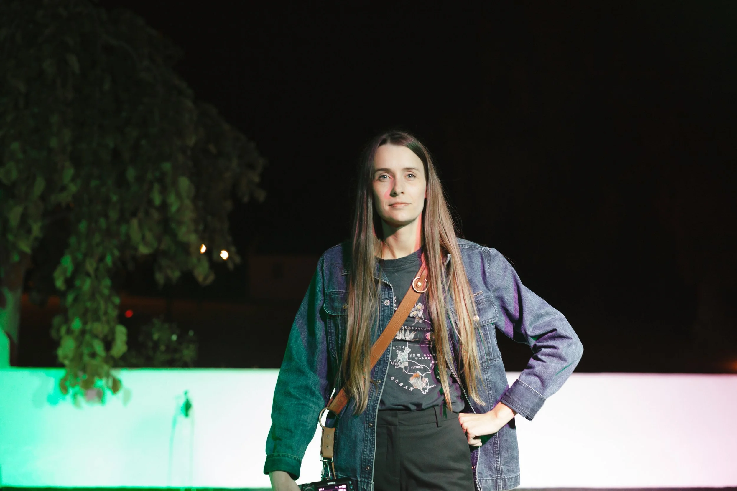 Young woman with long brown hair wearing a denim jacket, black t-shirt, and a tan crossbody bag standing outdoors at night near a tree and a green-lit wall. Gqeberha wedding photographer.