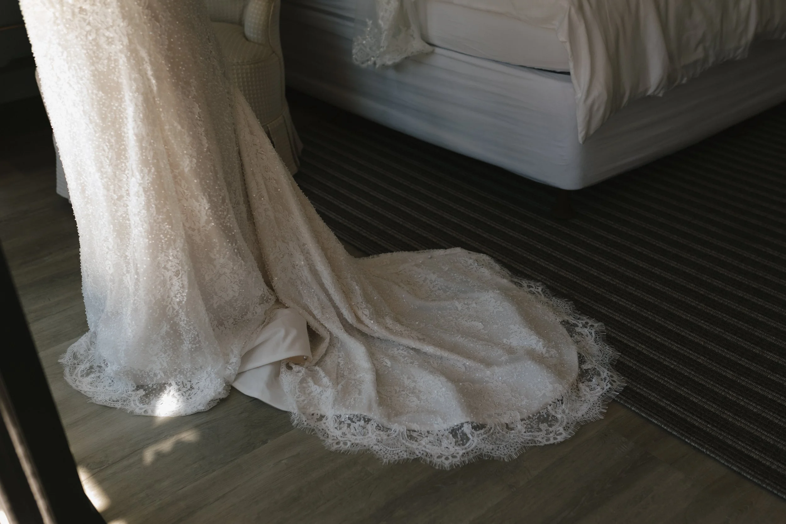 Wedding dress with lace details hanging over a bed in a softly lit room. Gqeberha wedding photographer at Royalston Lakehouse.