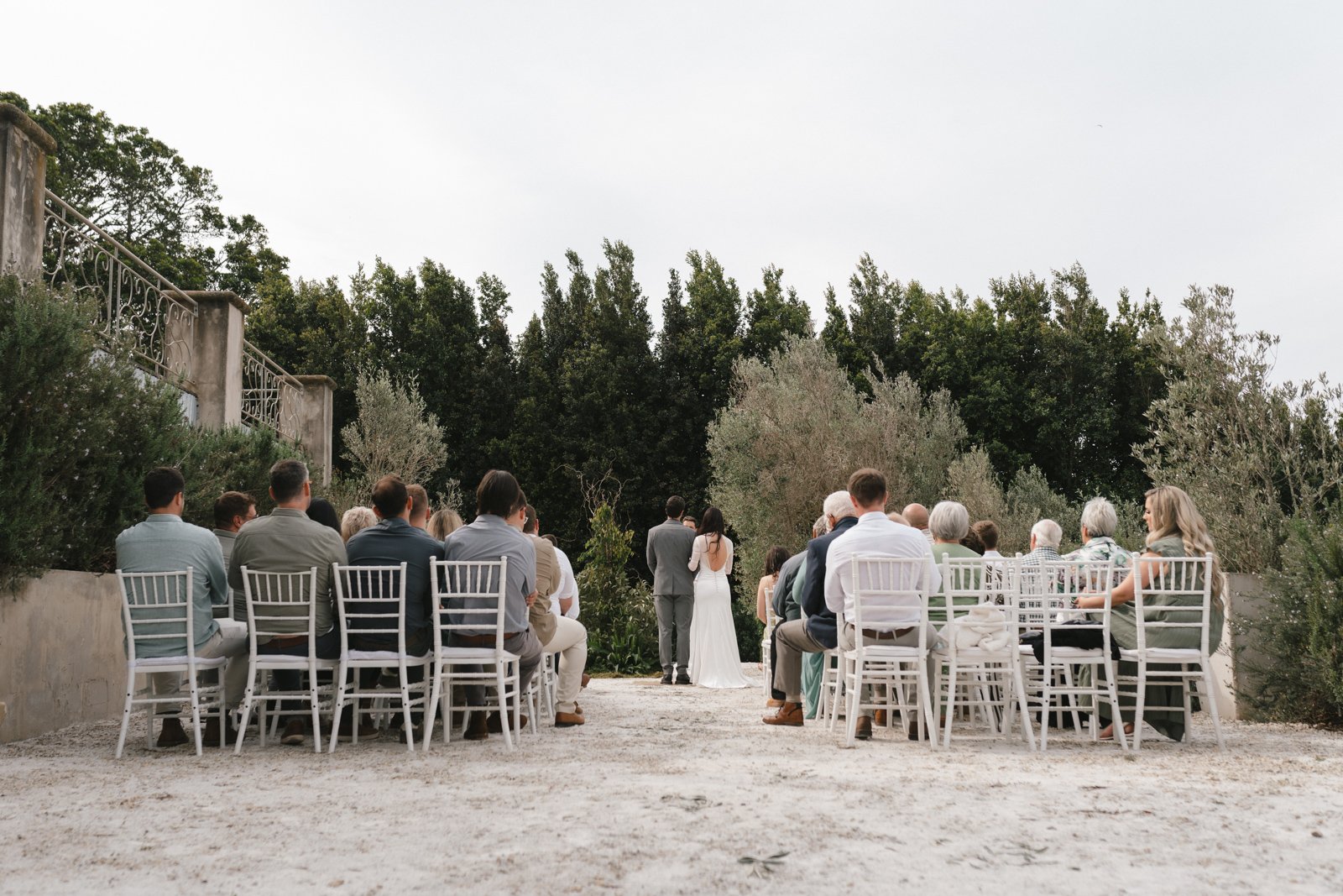 An outdoor wedding ceremony with guests seated on white chairs facing a bride and groom standing in front of a lush green backdrop.