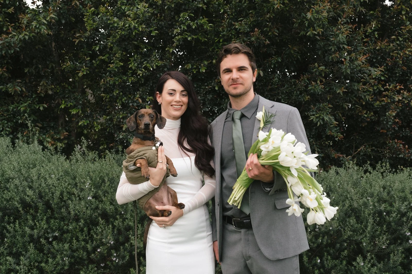 A couple dressed in wedding attire standing outdoors with shrubs and trees in the background. The woman is holding a small dog, and the man is holding a bouquet of white lilies.  Gqeberha wedding photography at The Rose Barn, Jefferys Bay