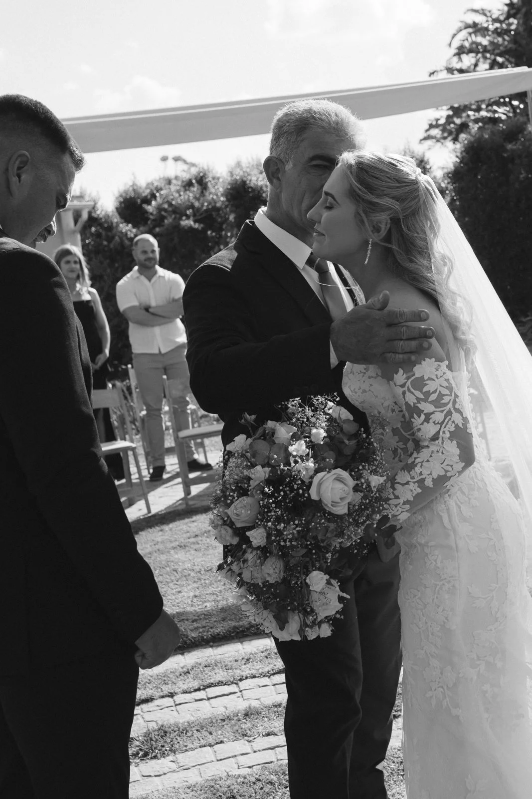 A bride and her father, share a tender moment during a wedding ceremony, with the bride holding a bouquet of roses and the man gently touching her face, surrounded by guests outdoors. Gqeberha wedding photographer at Lombardini Wedding Venue.