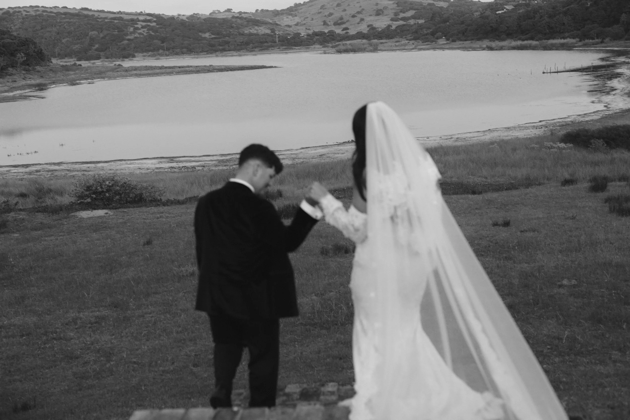 A couple dressed in formal wedding attire holding hands and walking away from the camera on a grassy area near a body of water, with hills and trees in the background, during blue hour. Royalston Lakehouse