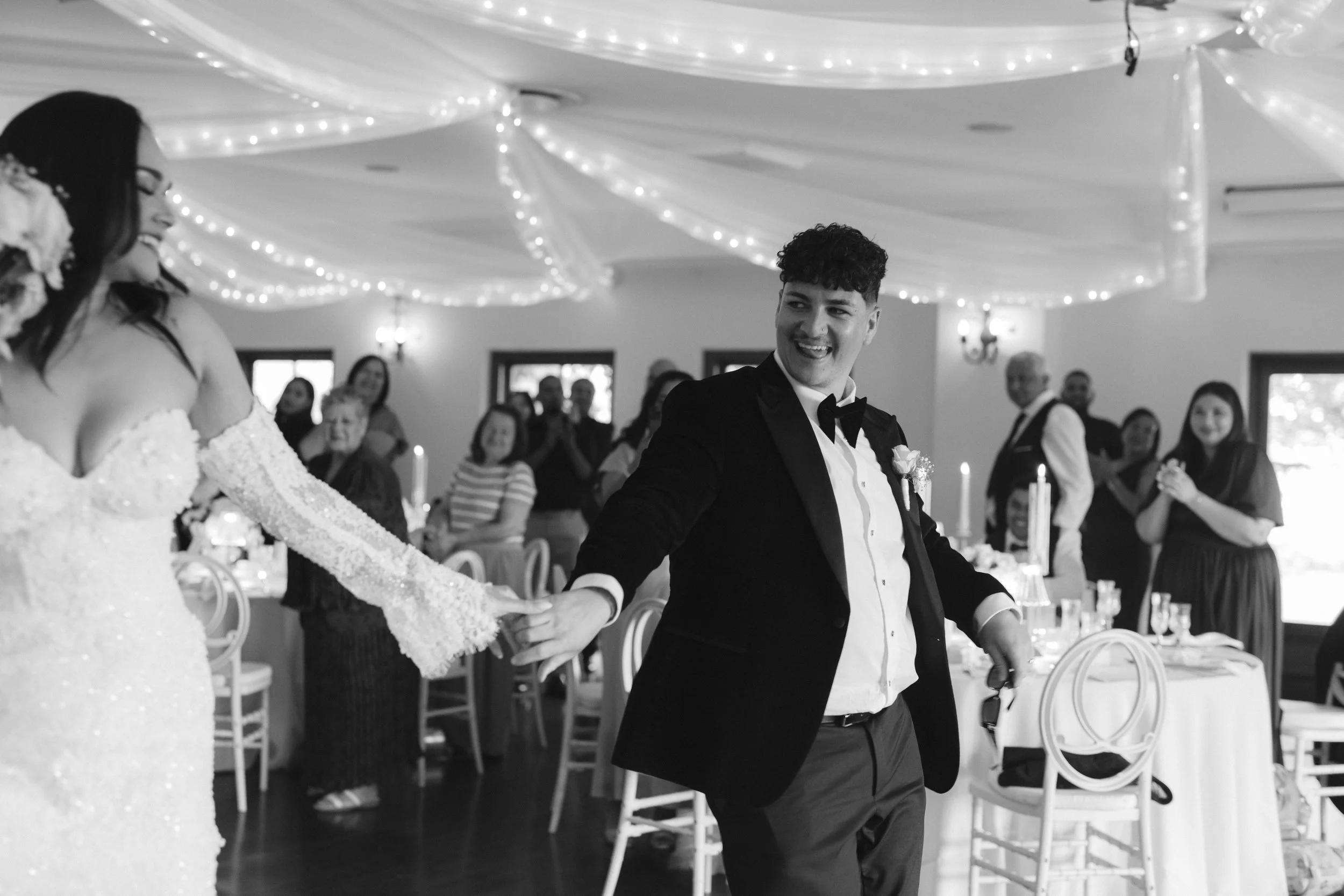 A groom and bride holding hands and dancing at their wedding reception in a decorated hall with string lights on the ceiling and guests watching. Gqeberha wedding photographer at Royalston Lakehouse.
