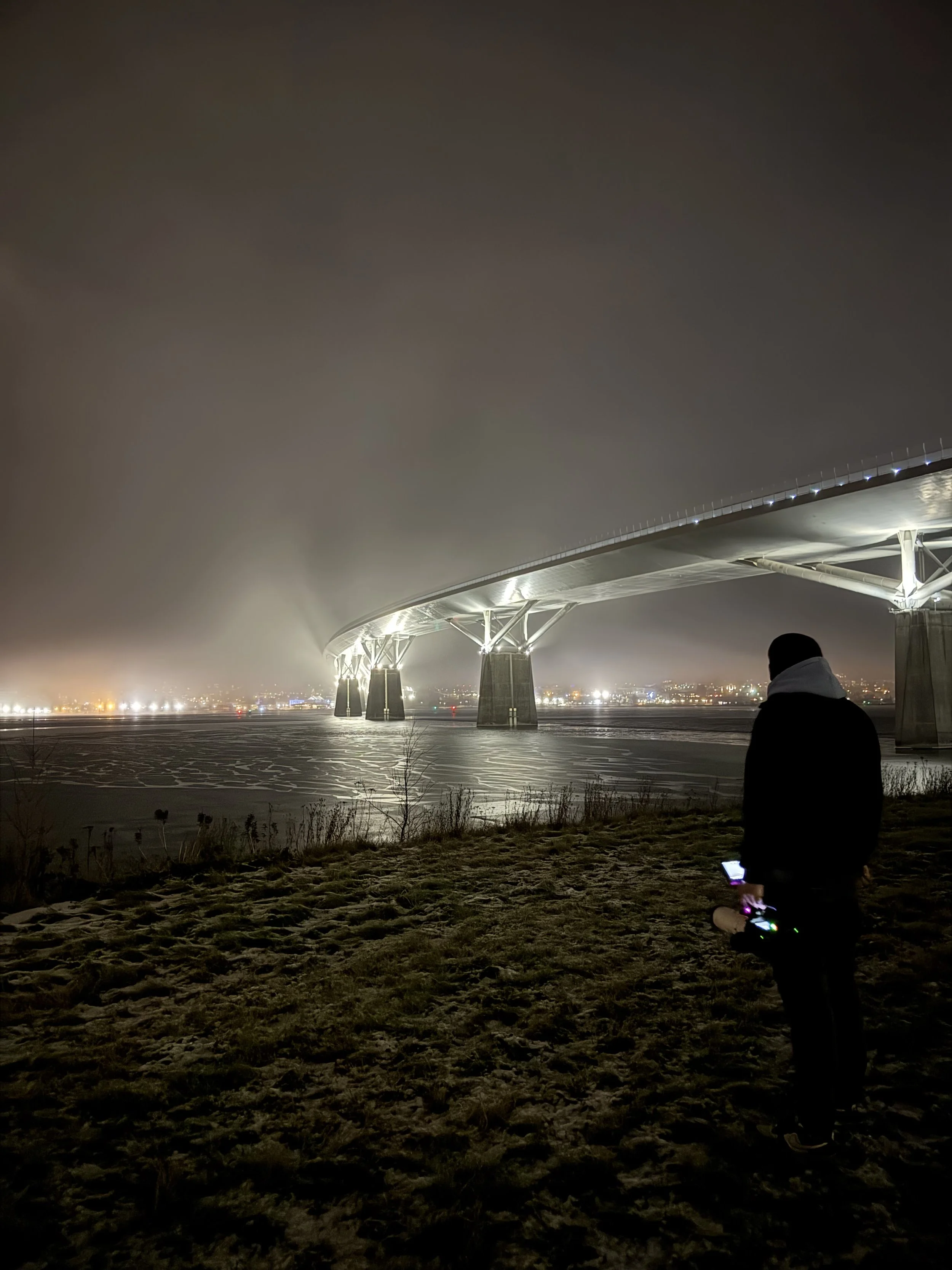 Nighttime view of a large illuminated bridge with multiple support pillars, a person standing on the ground near the river holding a device, and city lights in the background.