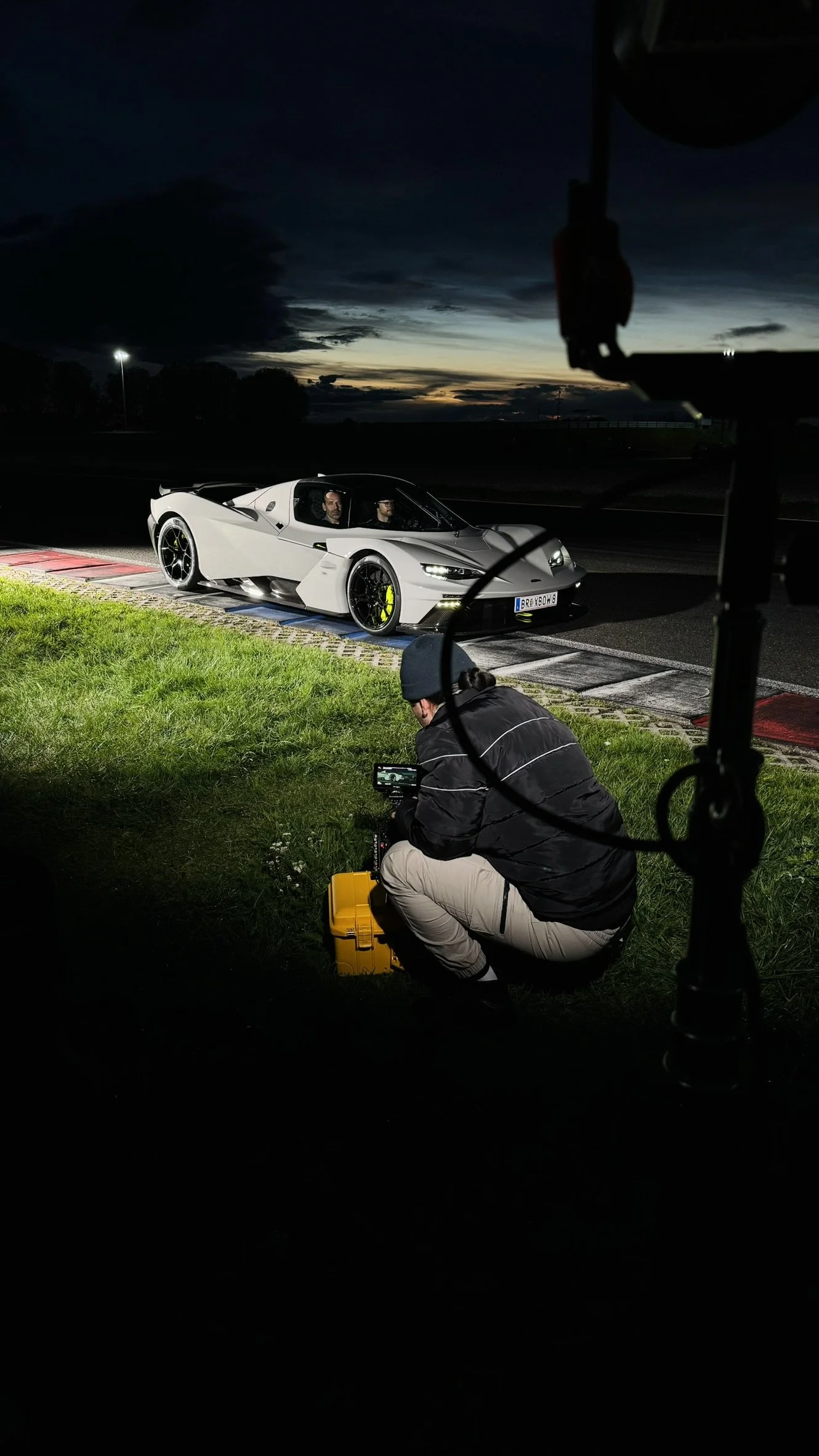 Nighttime scene of a white sports car on a racetrack, with a crew member kneeling on grass nearby, recording the car with a camera. The sky is dark with a hint of sunset on the horizon.