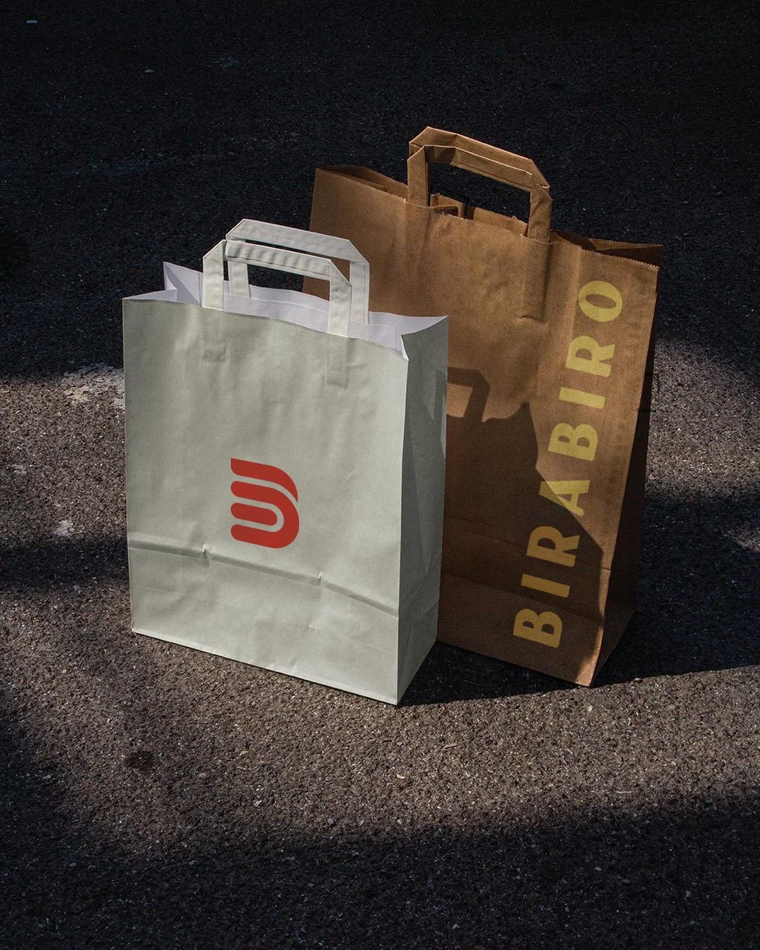 Two paper shopping bags, one white with a red logo and one brown with yellow text, standing on a dark asphalt surface.