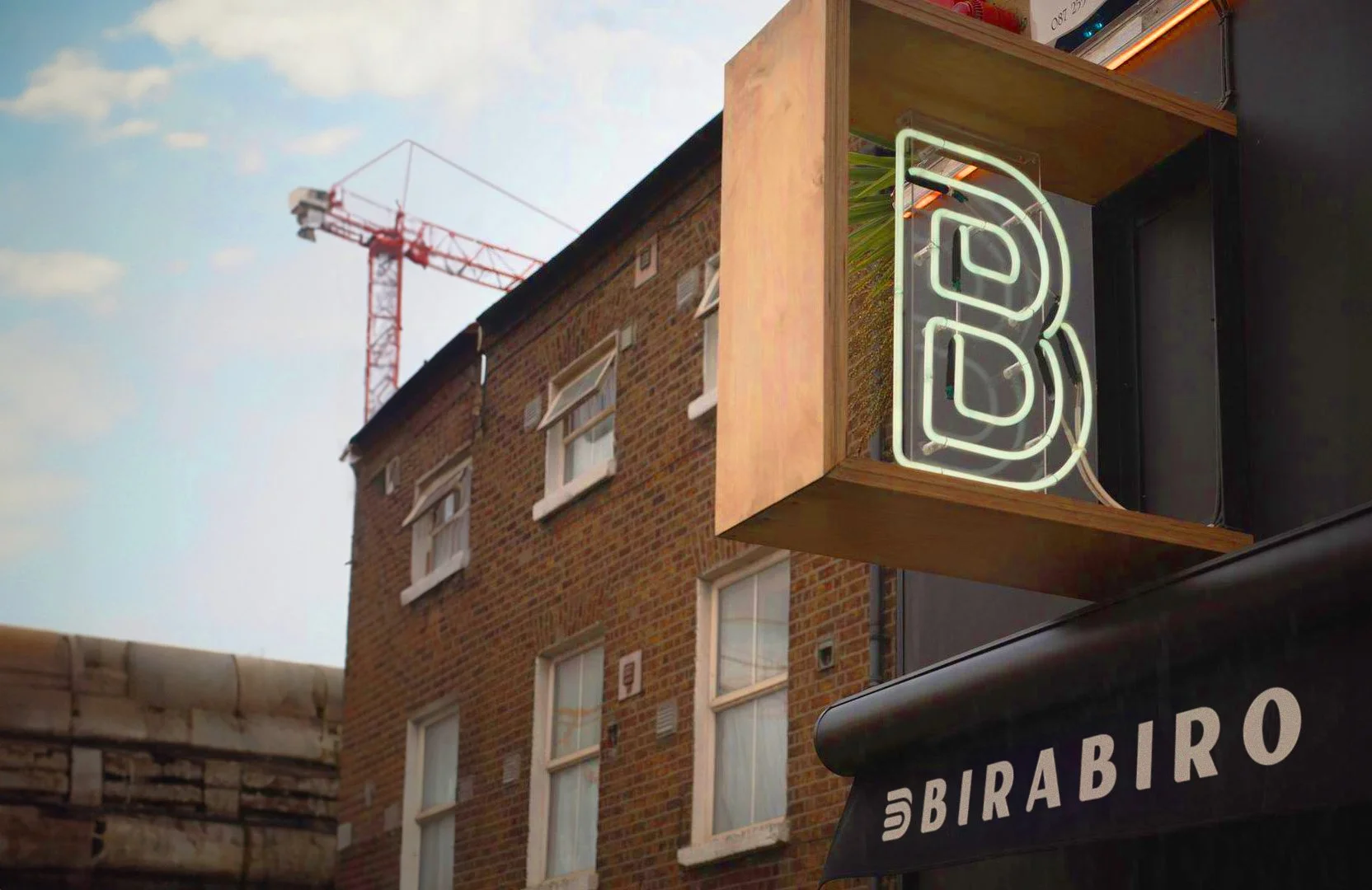 Neon sign that reads 'BIRABIRO' mounted on a wooden panel outside a building with brick walls and multiple windows, with a construction crane visible in the background against a blue sky with some clouds.