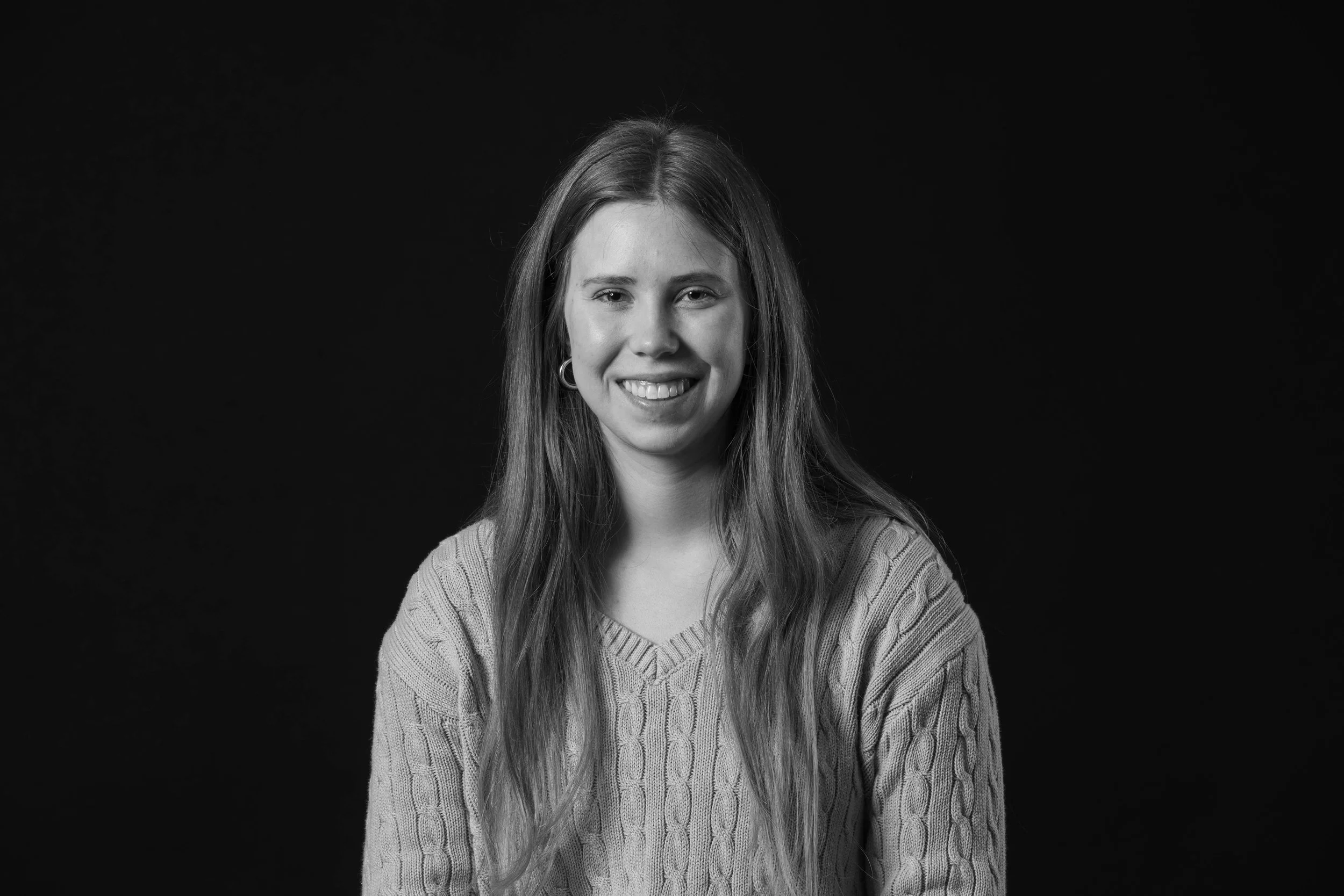 A young woman with long hair smiling, wearing a knit sweater, against a black background.