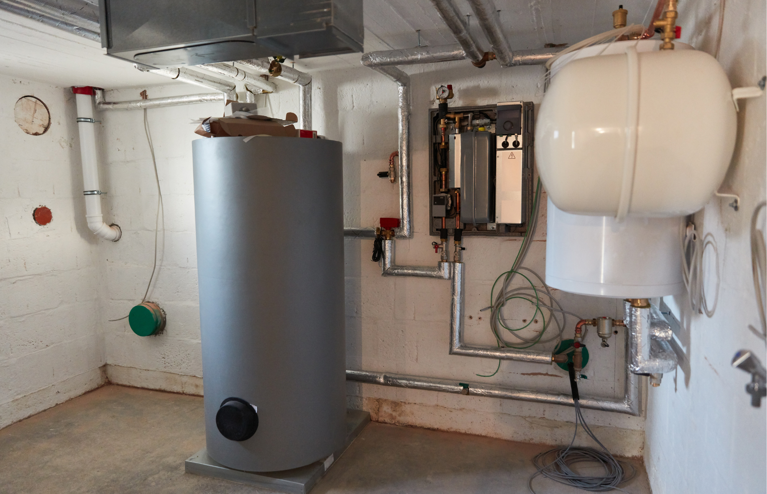 A basement utility room housing a hot water heater and a boiler system with associated pipes, valves, and electrical components.