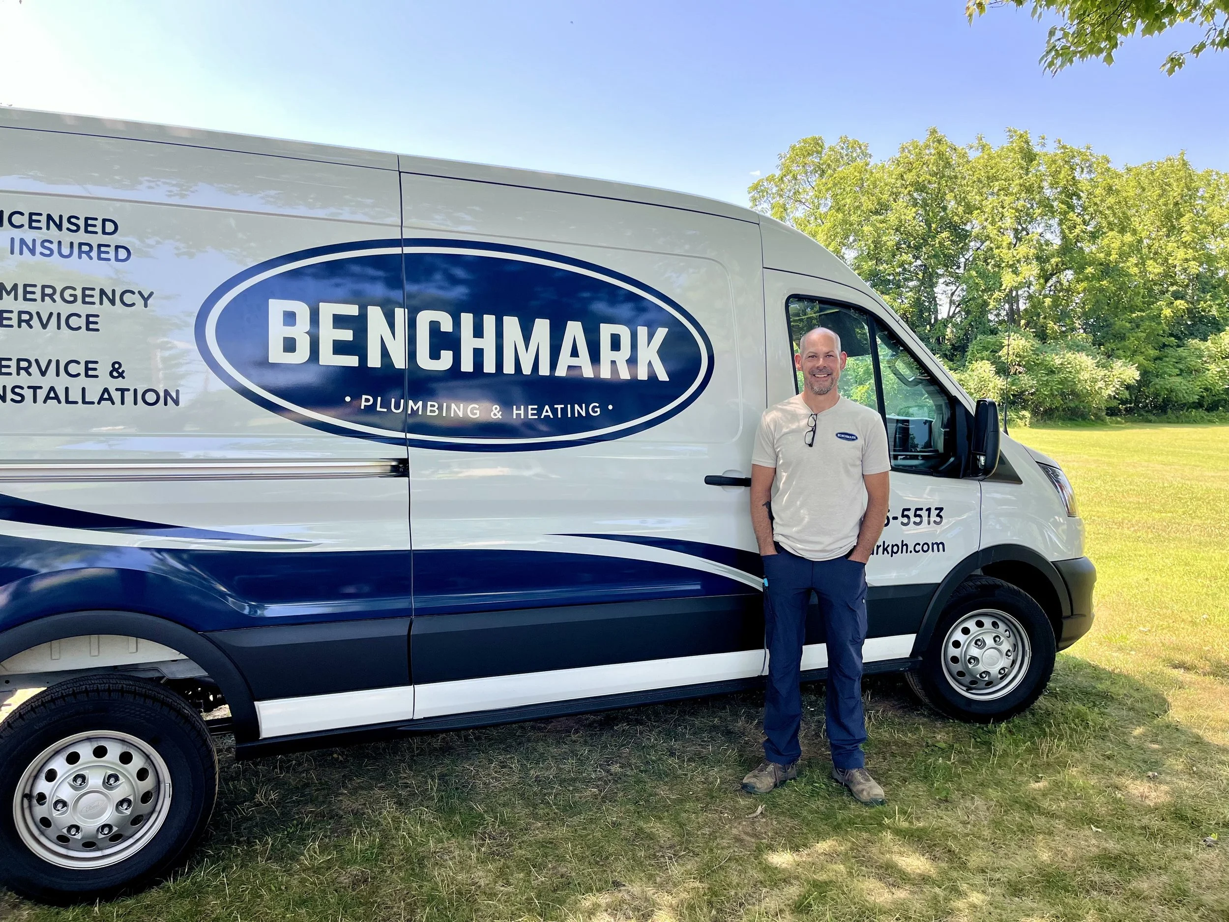 A man standing next to a white service van with the branding 'Benchmark Plumbing & Heating' in a park with green trees and a clear blue sky.
