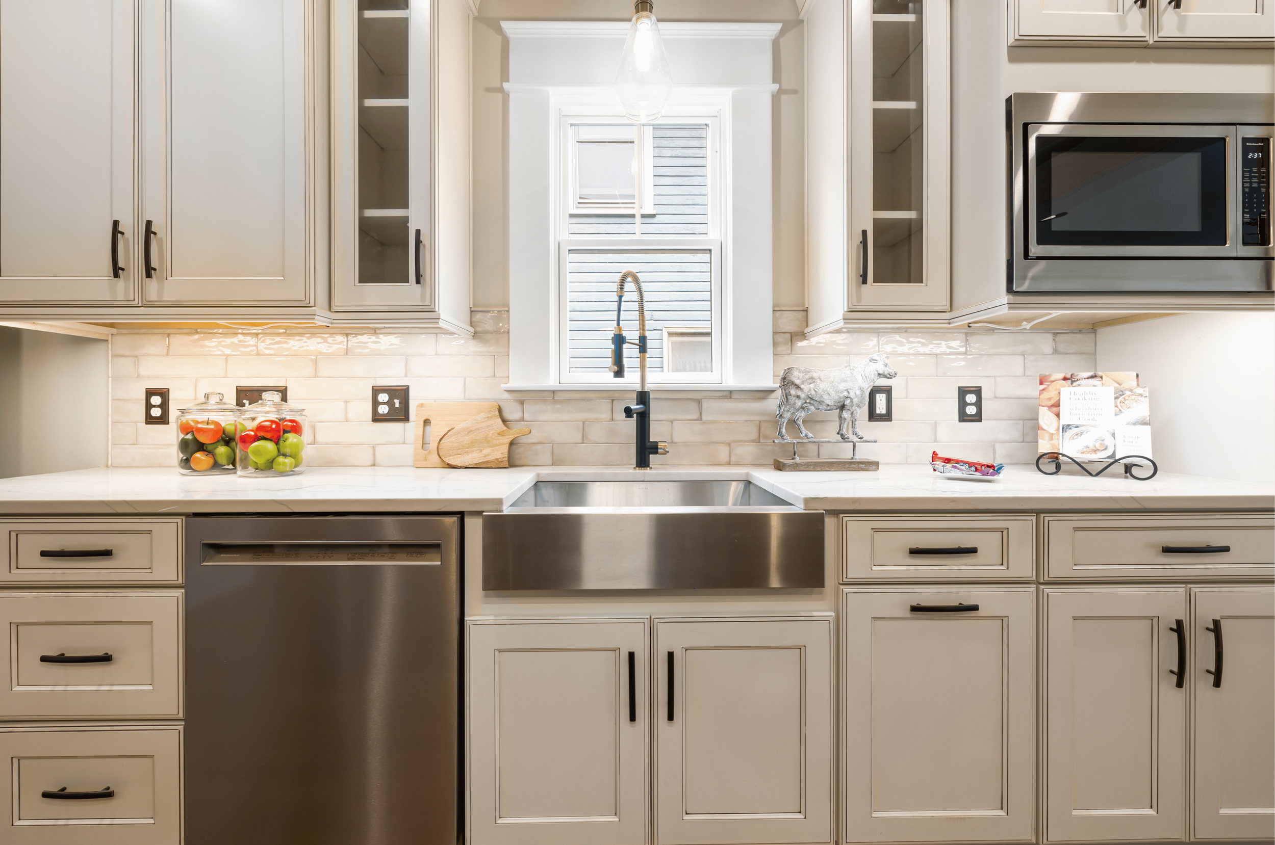 A modern kitchen with off-white cabinets, a brick backsplash, and a large window above the sink. The countertop has a jar of colorful apples, a wooden cutting board, and a decorative metal bull figurine. To the right, a built-in microwave oven is visible.