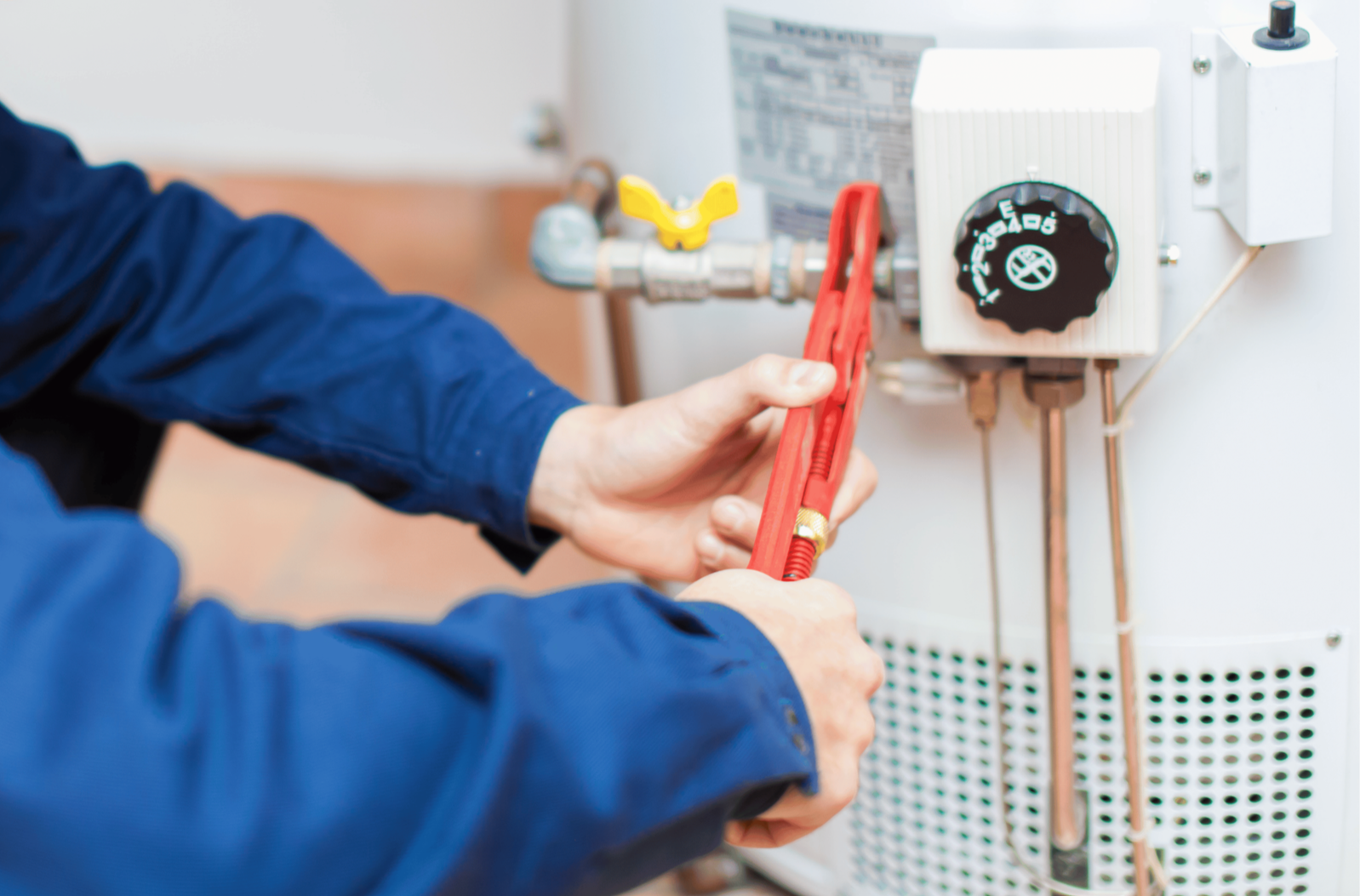 A person in a blue shirt is using a red pipe wrench to adjust or fix plumbing on a white water heater or boiler with pipes and valves.