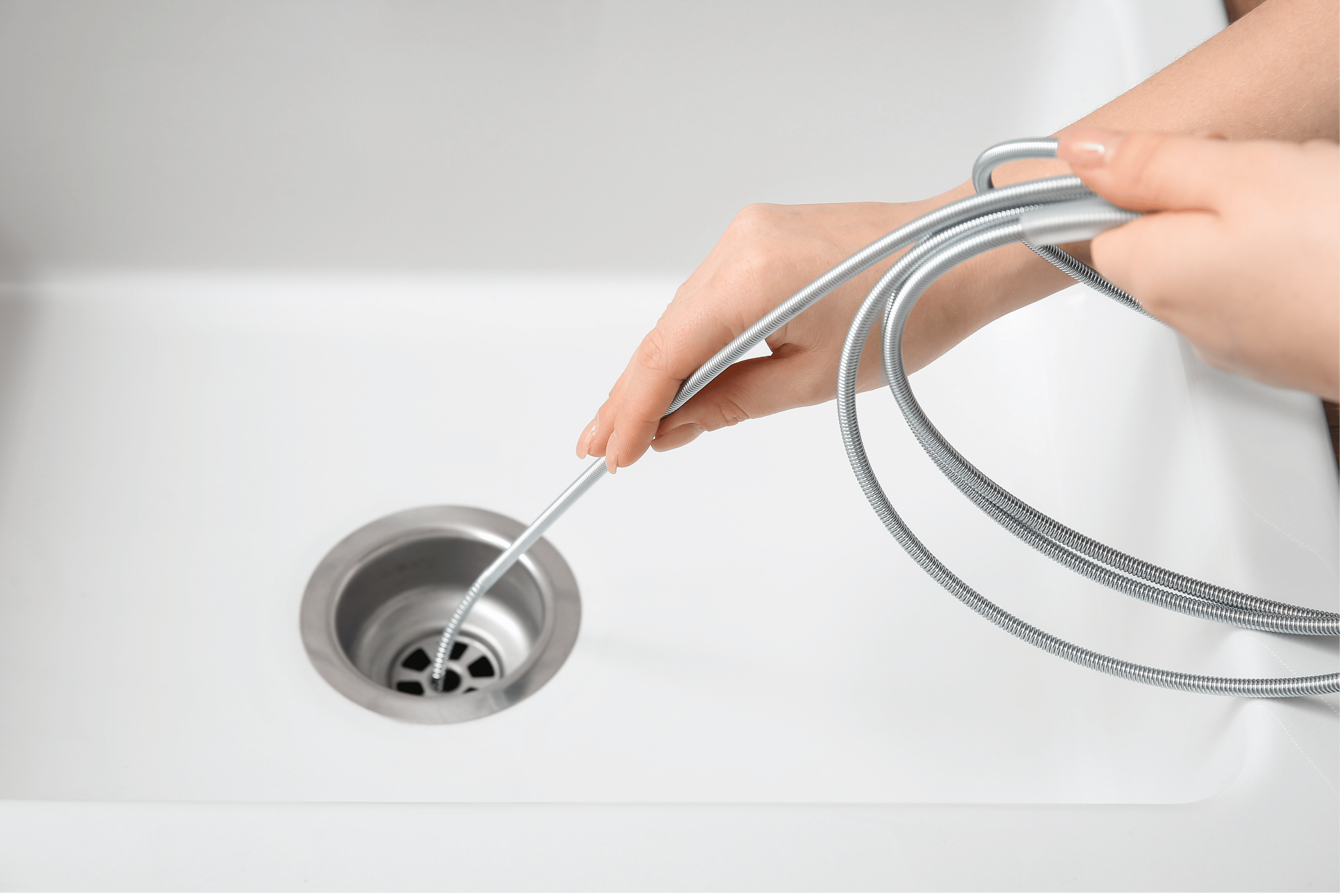 Person's hands holding silver flexible drain hoses over a white sink with a metal drain and a white background.
