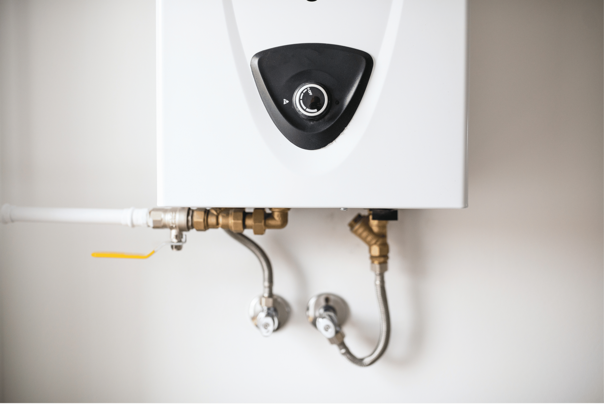 A white rectangular boiler with a black circular control knob in the center, connected to brass and silver pipes, mounted on a plain white and gray wall.