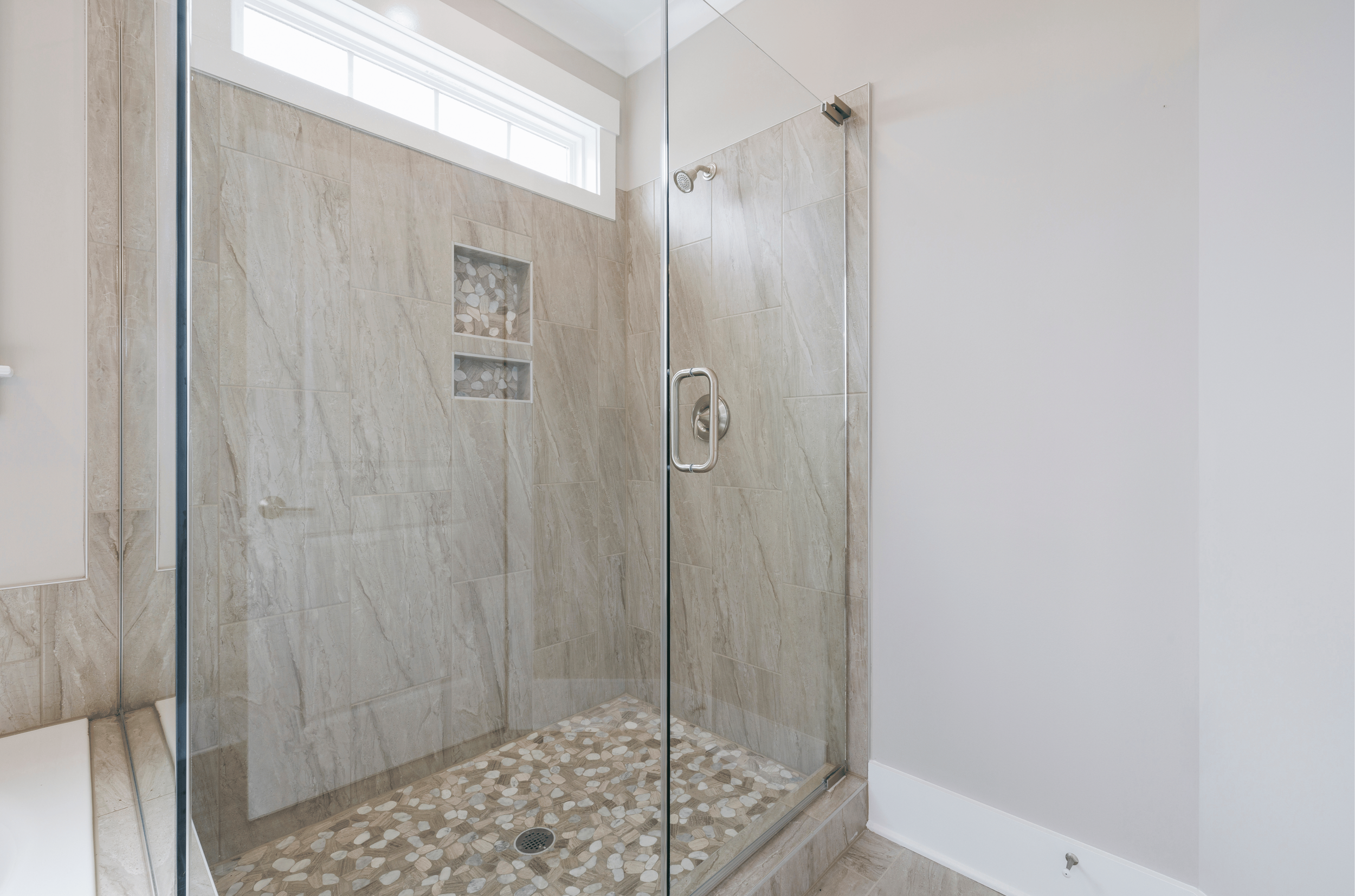 A modern shower with beige tile walls, a pebble floor, and a small niche with decorative stones, enclosed by clear glass doors, and a window above for natural light.