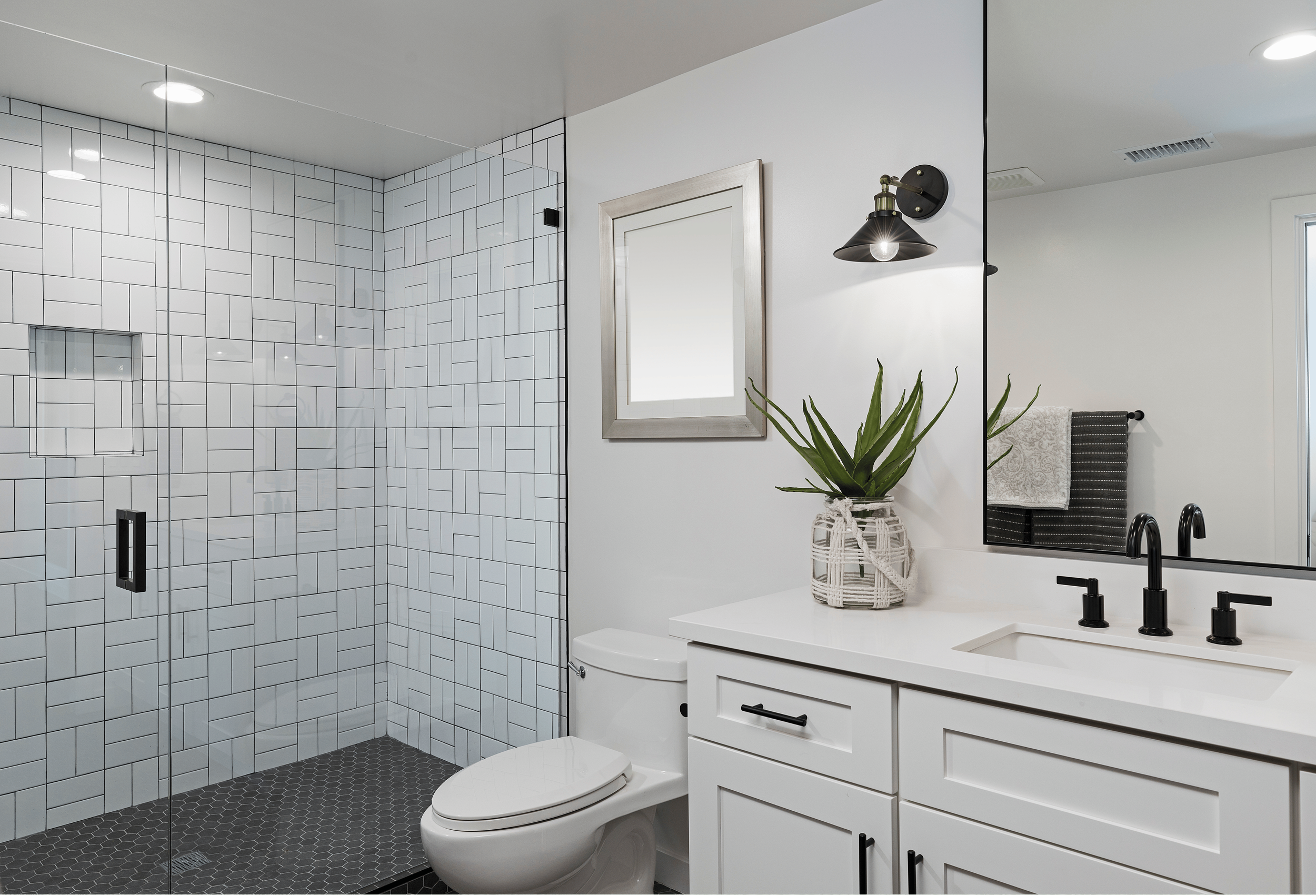 Modern bathroom with white and black decor, including a glass shower with white subway tiles, a white vanity with black faucet, and a mirror with a black frame. There is a potted agave plant on the vanity, a towel rack with towels, and wall-mounted light fixtures.