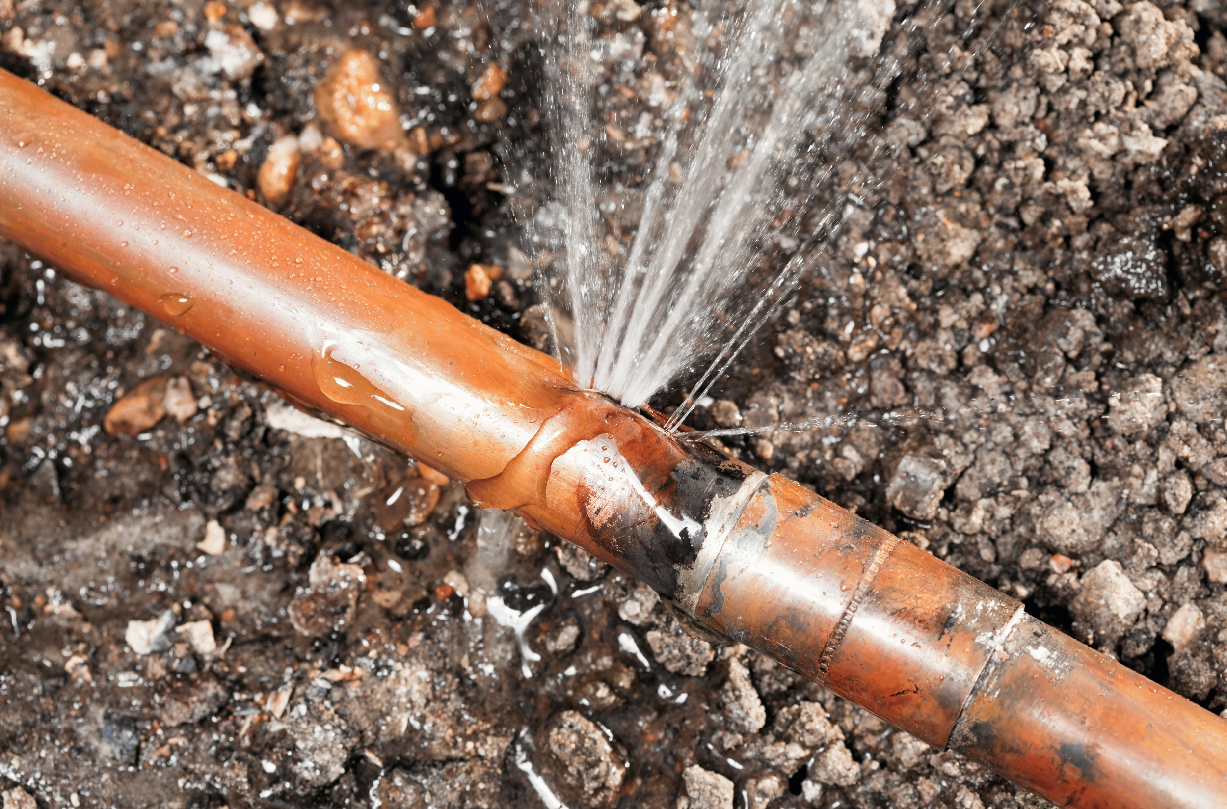 A close-up of an orange metal pipe lying on dark soil, with water spraying out of a crack in the pipe.