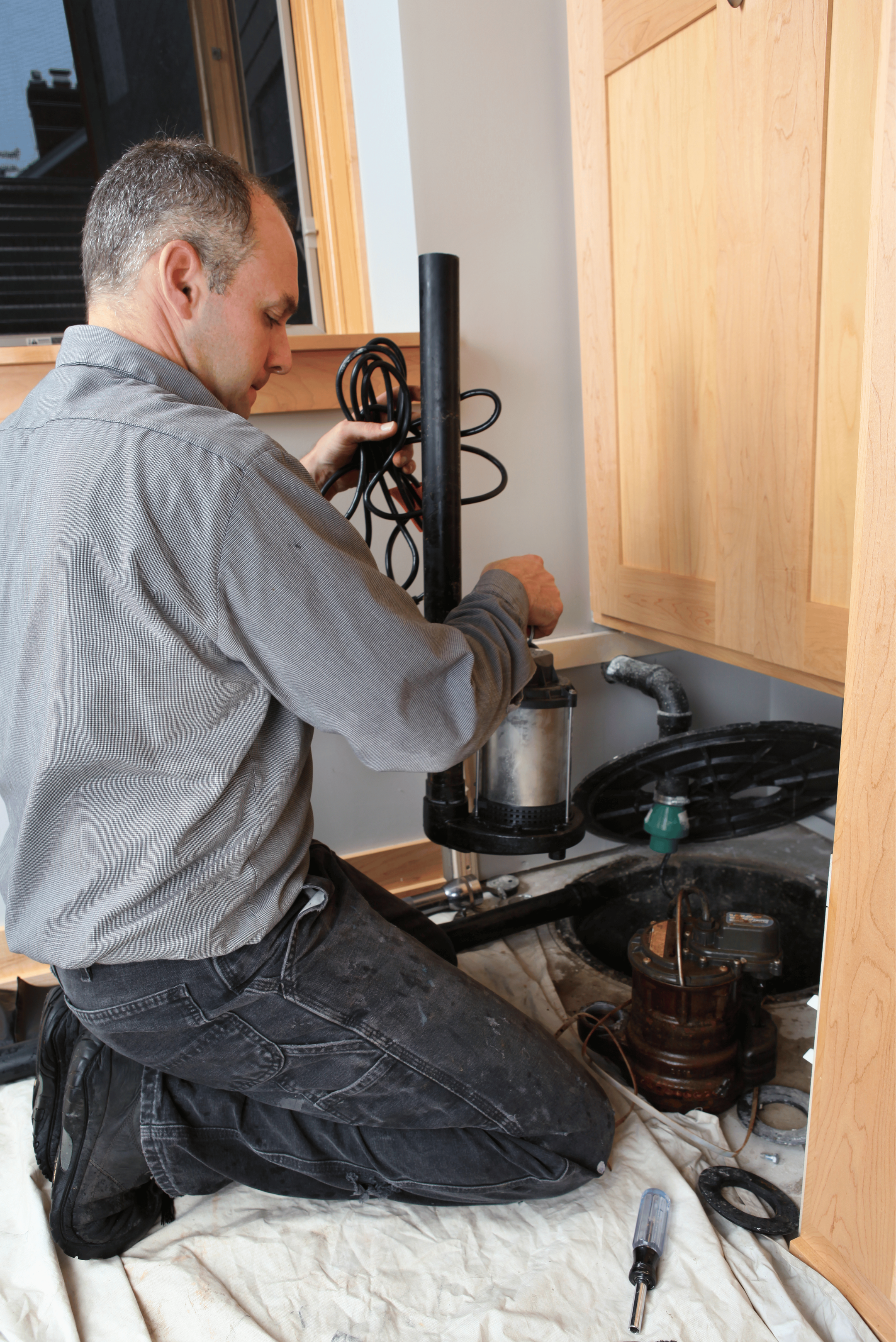 A man kneeling on the floor working on plumbing under a kitchen sink, with tools and pipes around him.