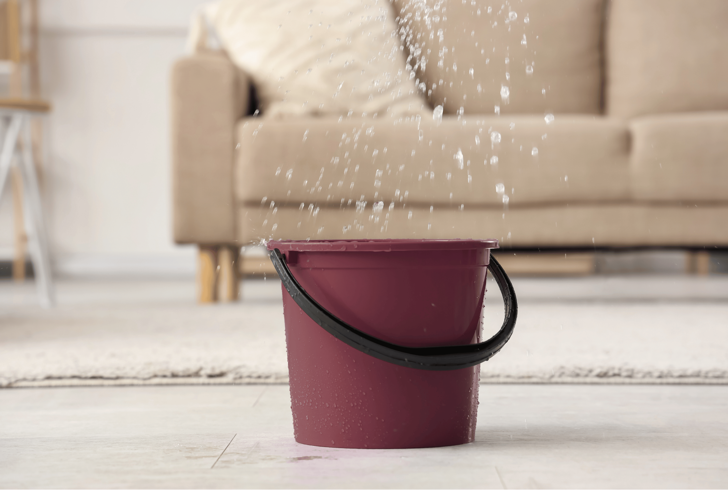 A pink mop bucket spilling water onto a light-colored wooden floor in a room with beige furniture.