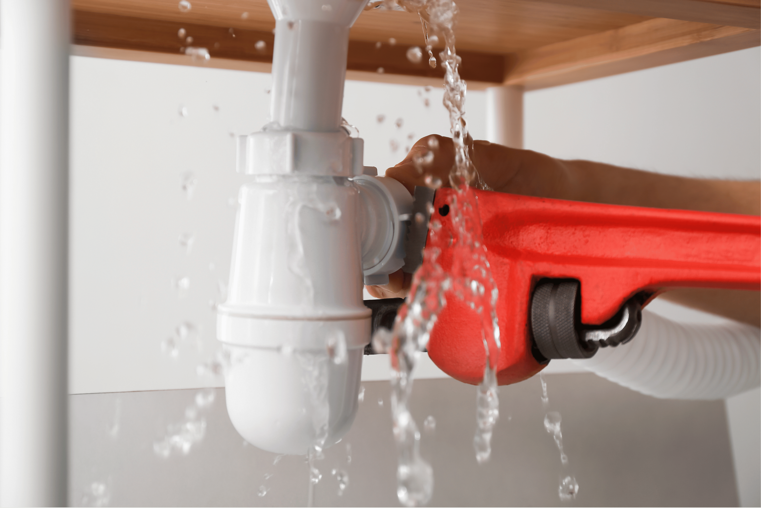 A person wearing a red tool belt is using a wrench to tighten a white PVC pipe fitting under a wooden table, with water flowing from the pipe.