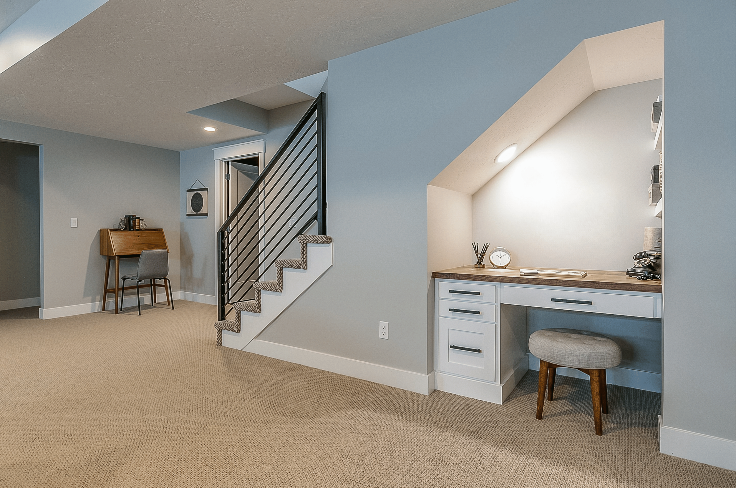 Interior of a home with beige carpet, light blue walls, a staircase with chevron patterned carpet, a small desk with a gray chair, and built-in desk area with a clock, pencils, and a telephone on the counter.