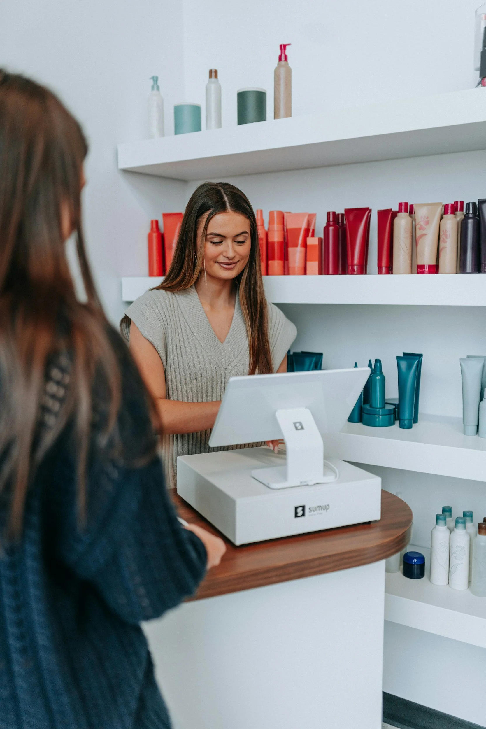 A woman at a counter takes an order from a customer at a retail or cosmetic store. The woman is smiling and looking down, with shelves of colorful bottles and containers behind her.
