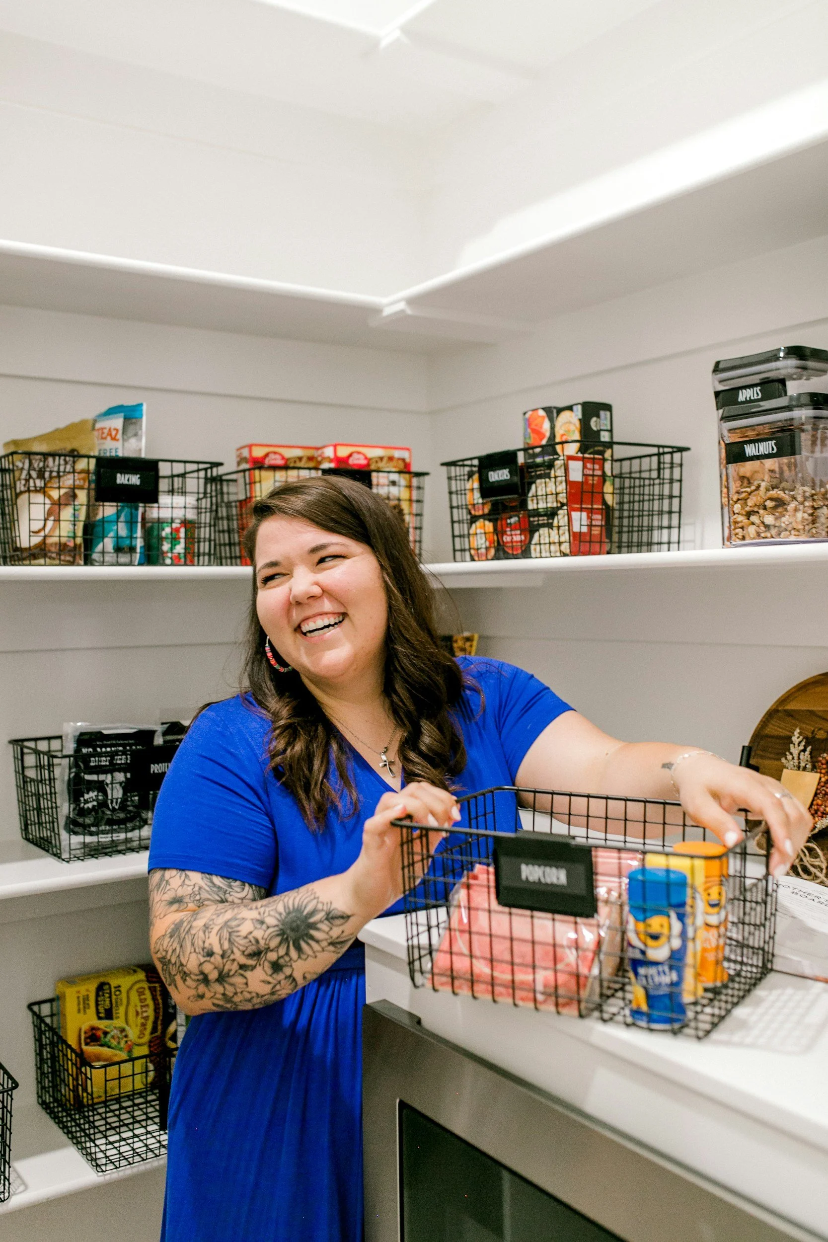 A woman with tattoos on her arms, wearing a blue dress, smiling and shopping at a store. She is standing in front of shelves filled with snacks and nuts, reaching into a basket labeled 'Nuts'.