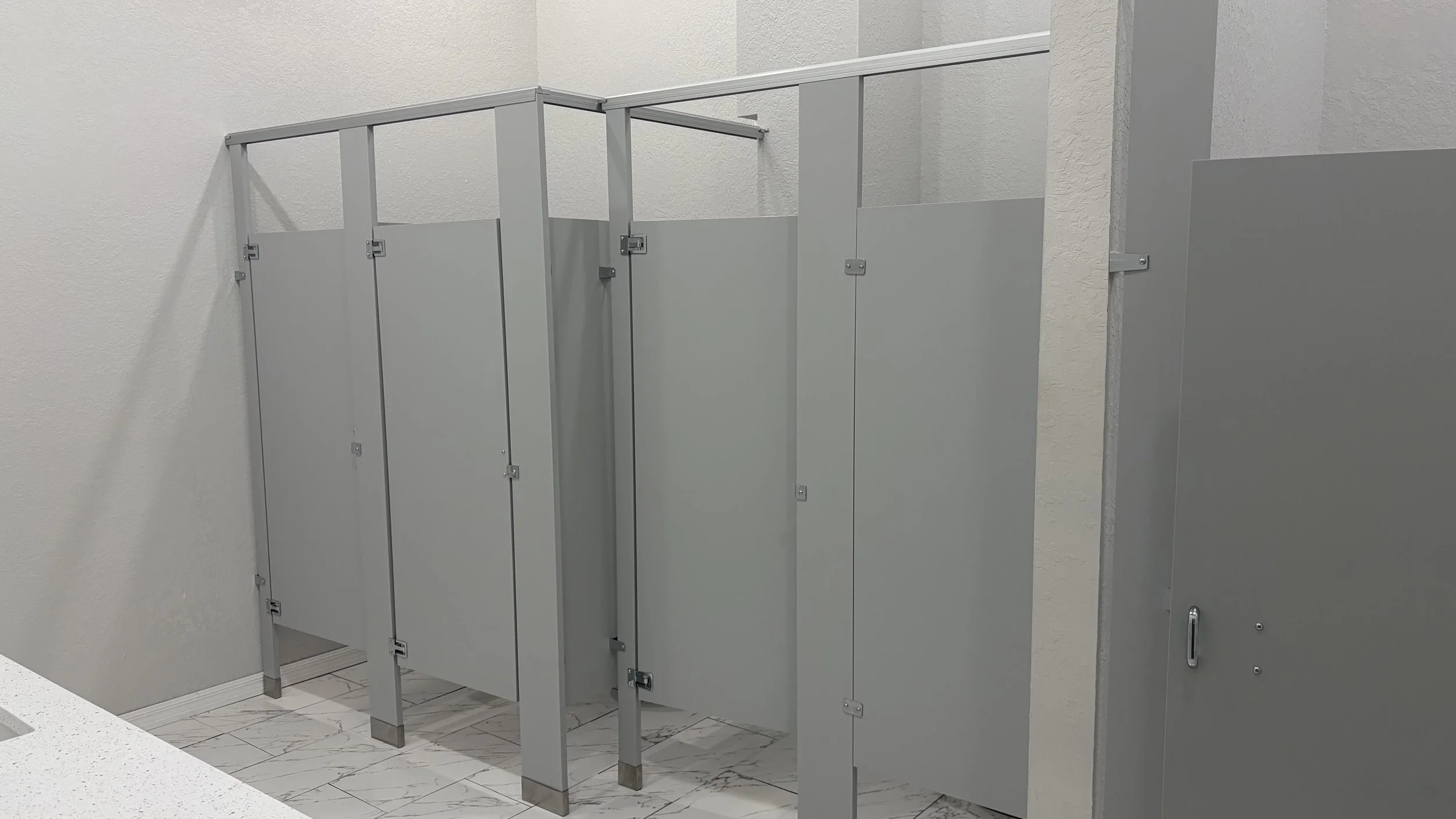 A row of grey public restroom stalls with closed doors, set against white textured walls and a marble-effect tiled floor.