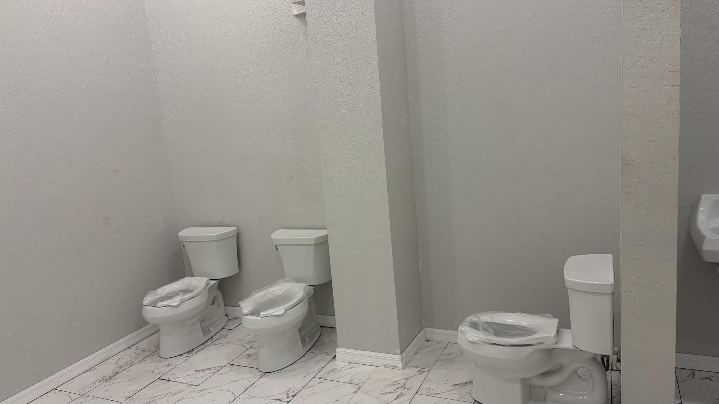 Public restroom with three toilets, two of which are side by side and one separate, all with plastic covers, and a small white sink on the right.