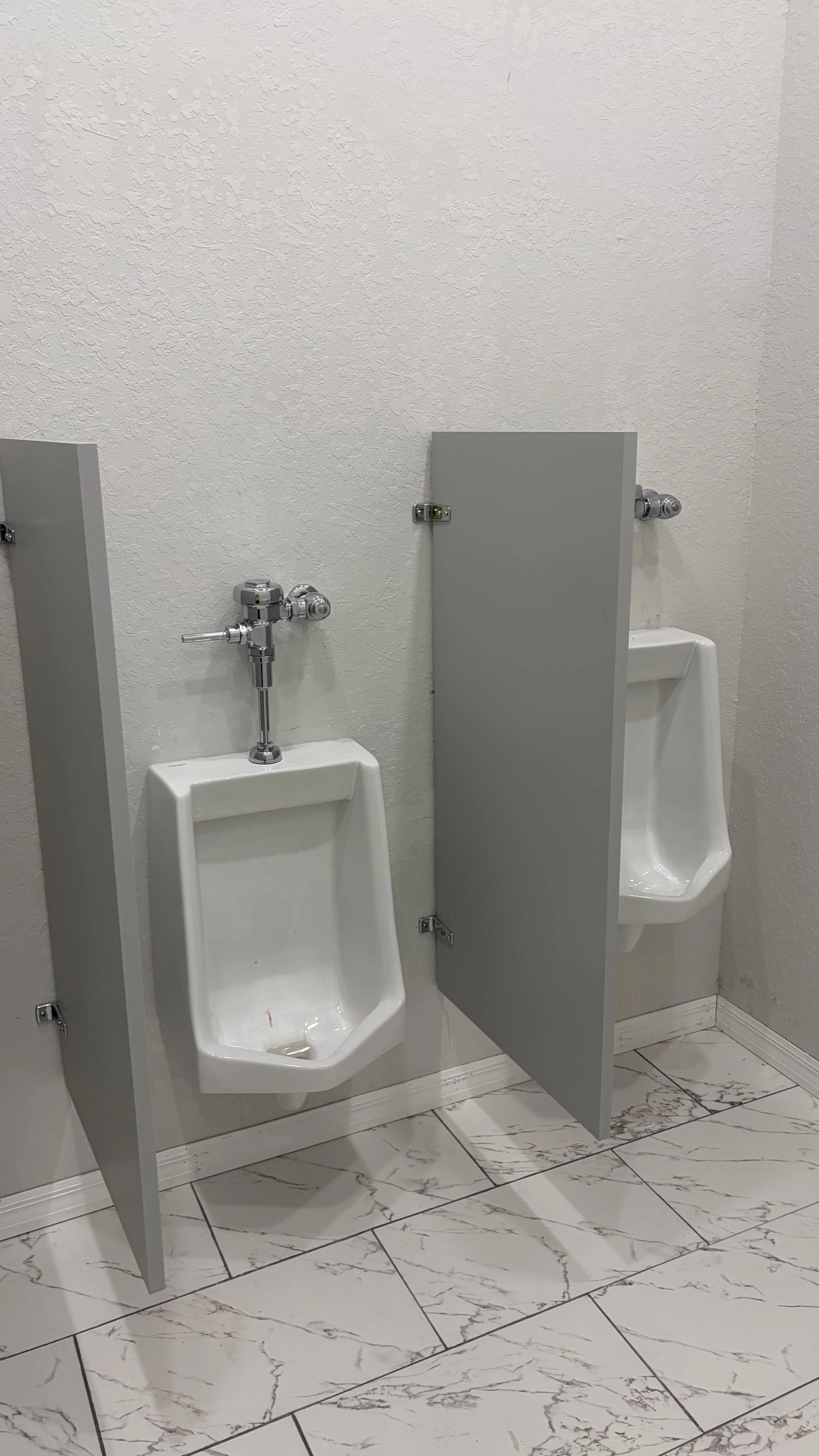 Two urinals separated by a gray privacy divider in a men's restroom with tiled floor and textured walls.