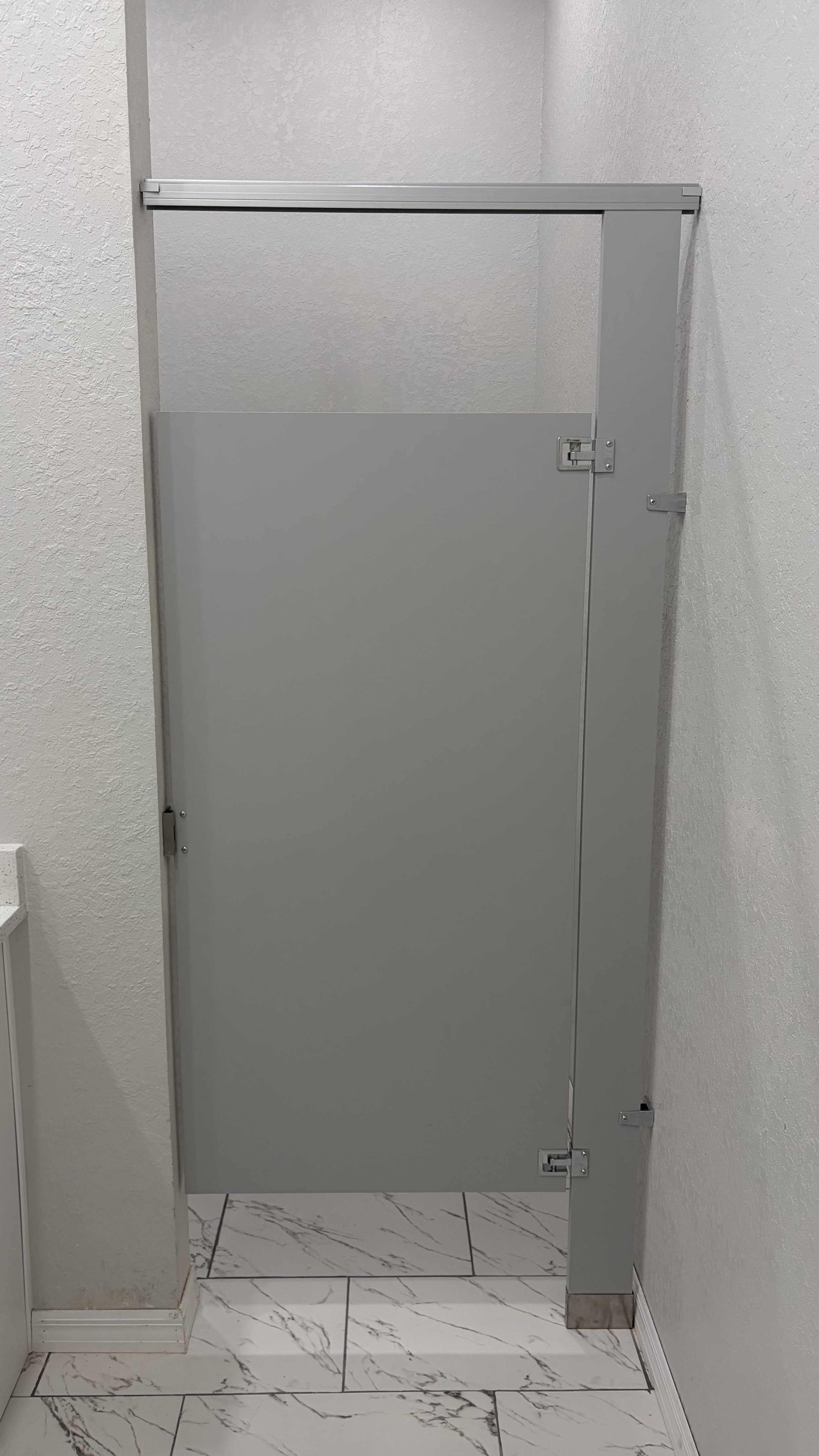 A gray bathroom or restroom stall door with a lock, on a bathroom with white marble floor tiles and white textured walls.