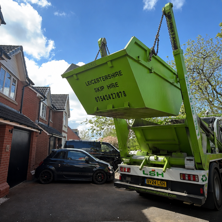 A green skip is being lifted by a large truck, with the skip displaying sign for Leicestershire Skip Hire, parked in front of residential houses with cars on the driveway under a blue sky with clouds.