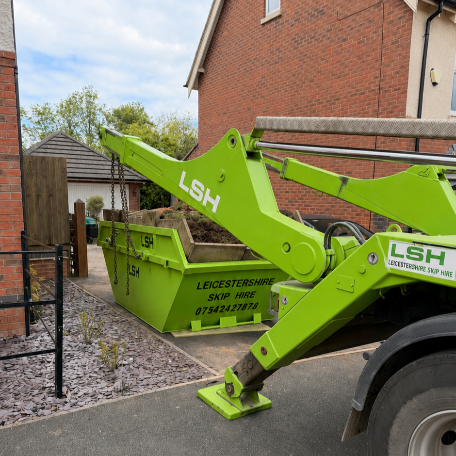 A green mini loader lifting a container of soil in a residential backyard. The container and loader are marked with 'LSH' and 'Leicestershire Skip Hire' along with a phone number.