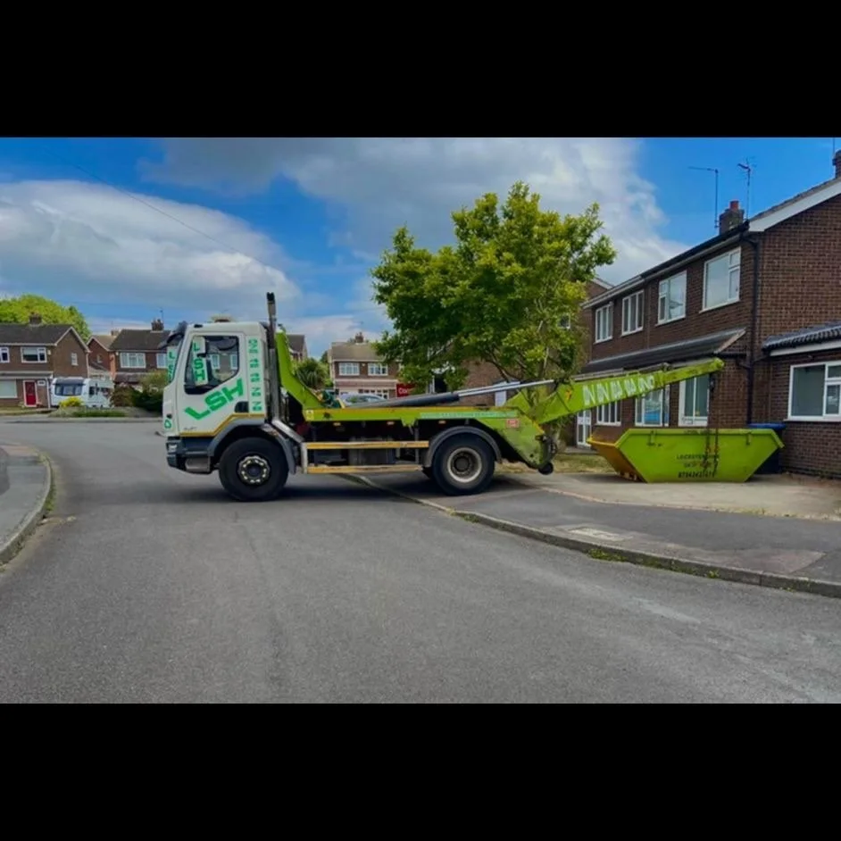 A green and white skip lorry parked on a residential street, with its boom extended to lift a dumpster in front of a brick house.