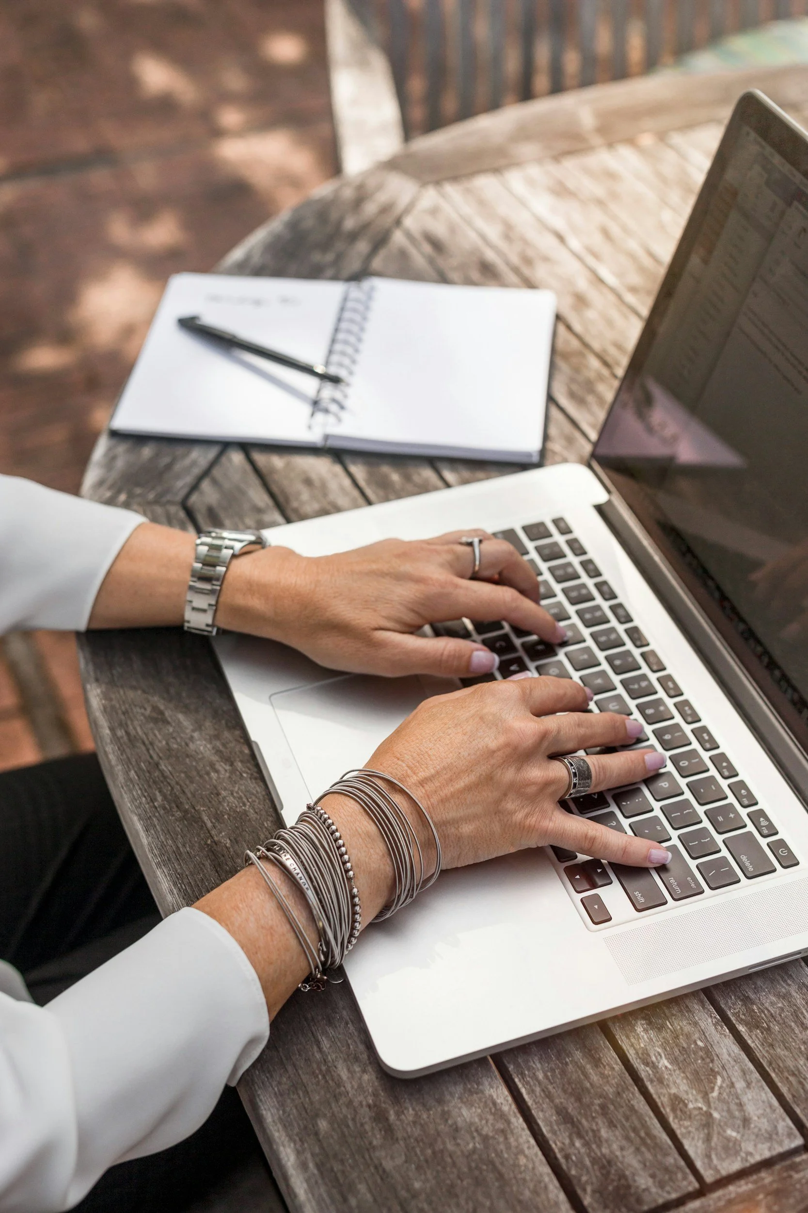 Close-up of a person typing on a white laptop with a silver watch and various bracelets on their wrist, placed on a wooden table outdoors. An open notebook with a pen rests on the table behind the laptop.