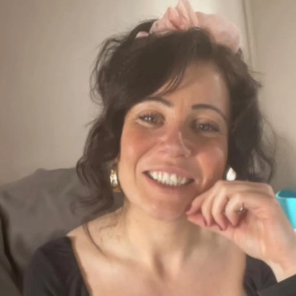 A woman with dark hair wearing a pink hair bow, smiling and resting her chin on her hand.