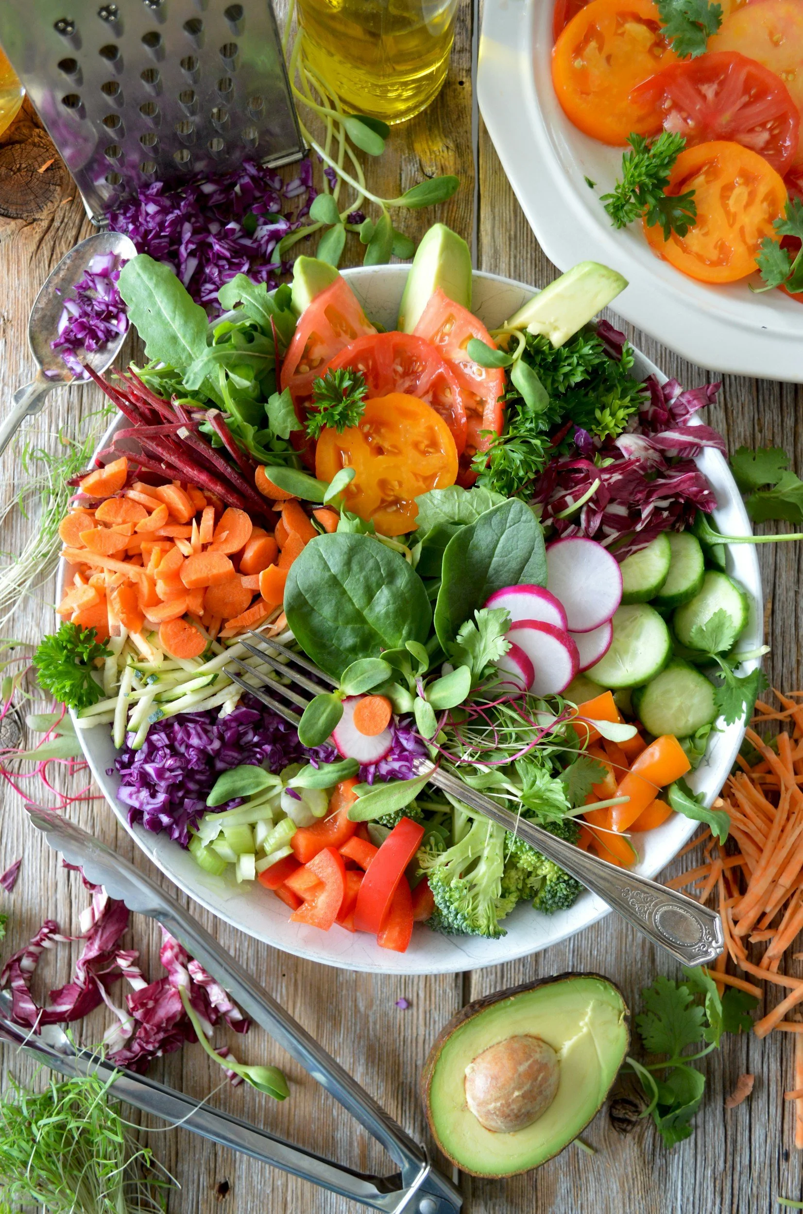 A large bowl of fresh vegetable salad with tomatoes, radishes, carrots, cucumbers, lettuce, and herbs on a rustic wooden table, with additional ingredients like grated carrots, avocado, and shredded purple cabbage nearby.