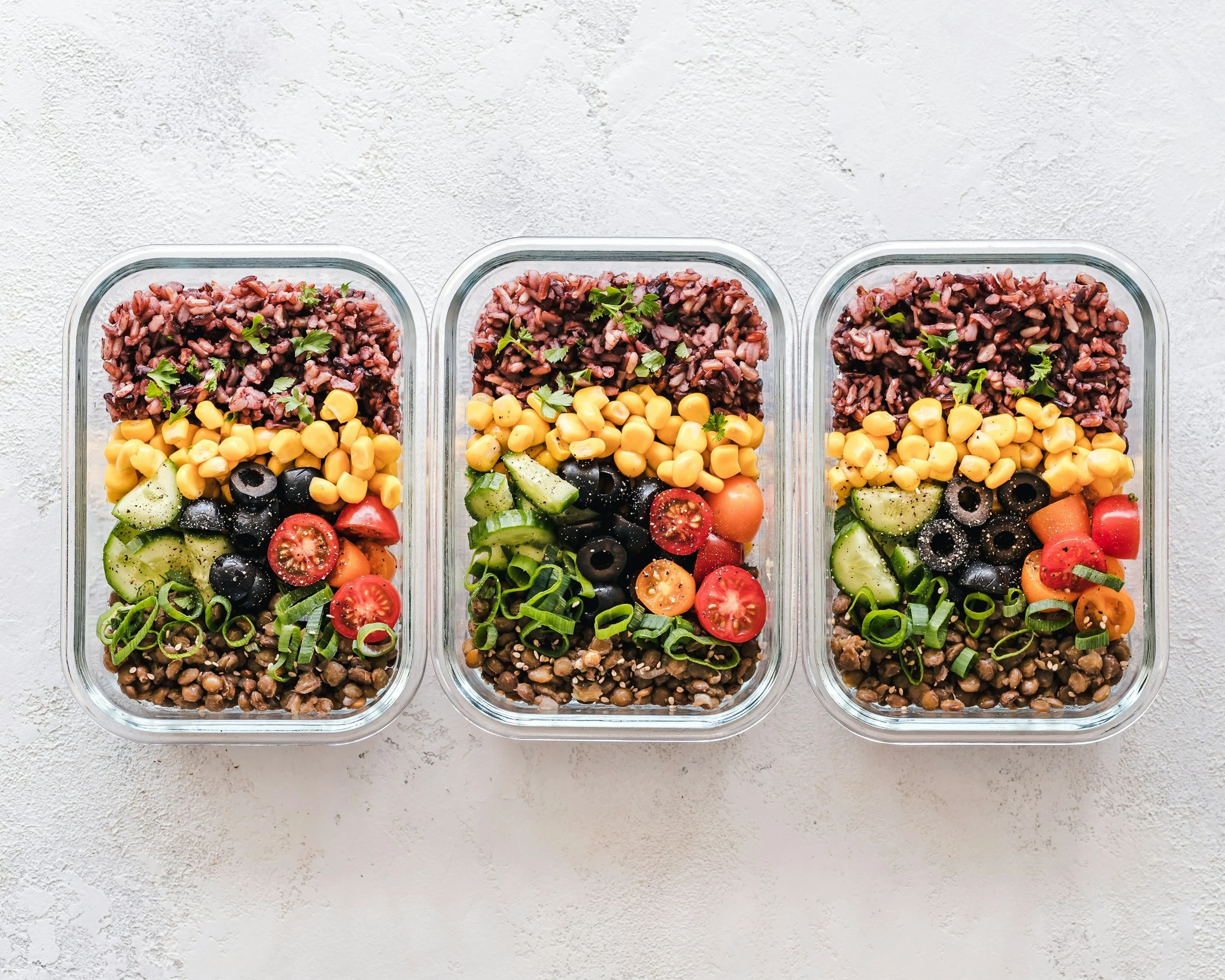 Three glass containers filled with salad ingredients such as cherry tomatoes, cucumber slices, black olives, corn, chopped green onions, and fresh herbs, placed on a light textured surface.