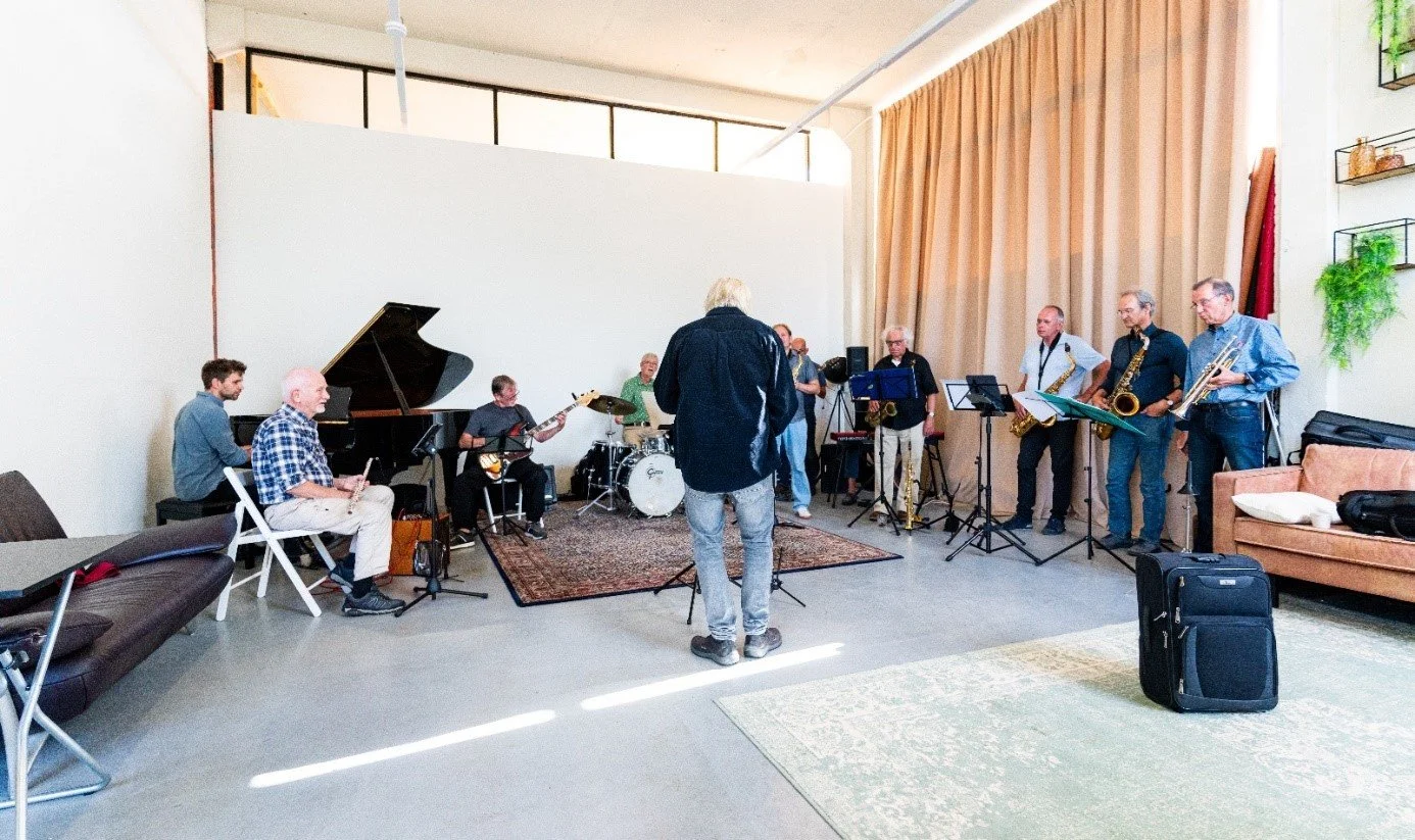 Muziekband speelt live in een woonkamer met een jazz-setup, inclusief pianospeler, gitarist, drummer en saxofonisten, terwijl een dirigent of bandleider voor hen staat.