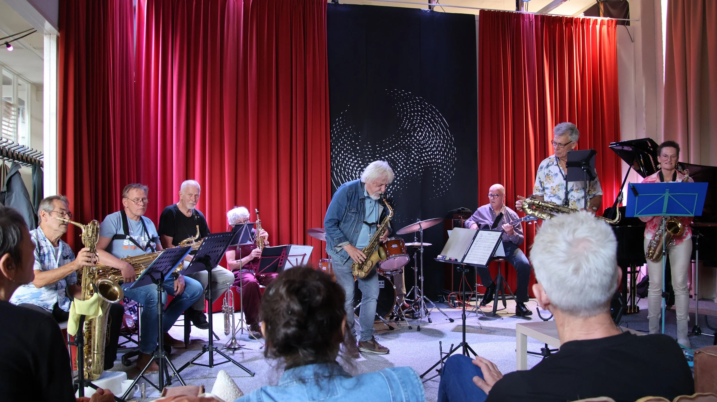 Een jazzband speelt op een podium met rode gordijnen, bestaande uit saxofonisten, gitaristen en een dirigent, terwijl het publiek ernaar kijkt.