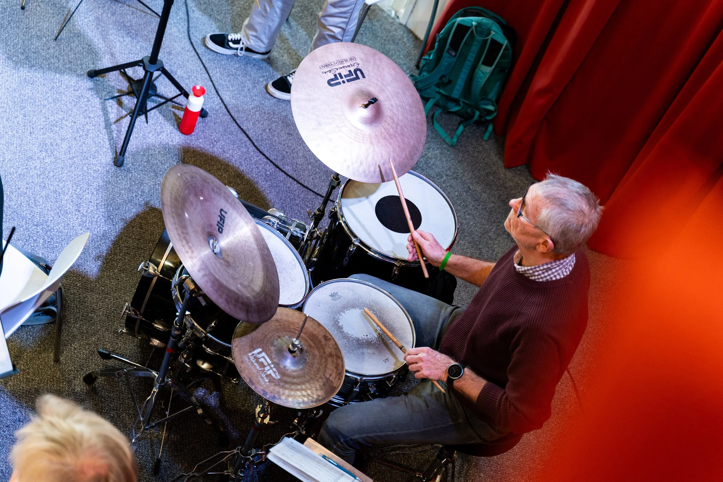 Een oudere man met grijs haar en een bril speelt drums in een zaal met een rode gordijnwand.