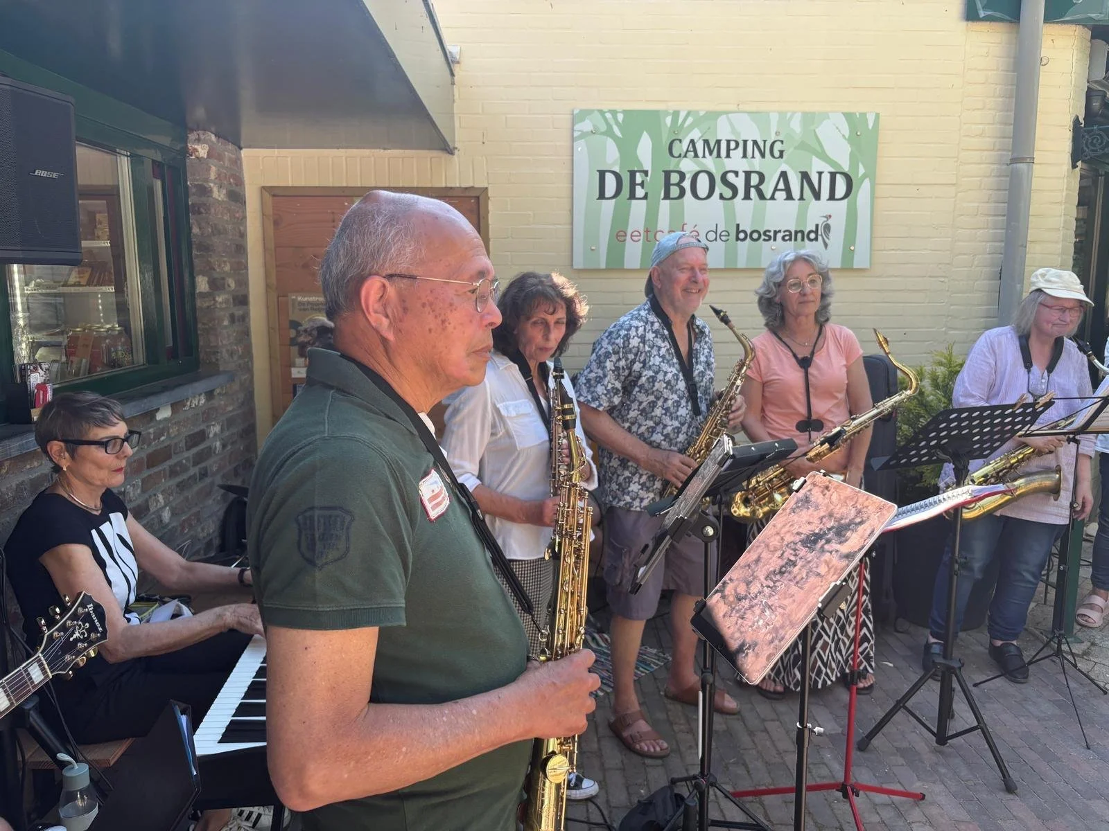 Een groep muzikanten speelt buiten voor een cafe genaamd 'Camping De Bosrand'. Ze bestaan uit verschillende mensen met instrumenten zoals saxofon, gitaar, keyboard en blaasinstrumenten, en ze lijken te genieten van een live optreden.