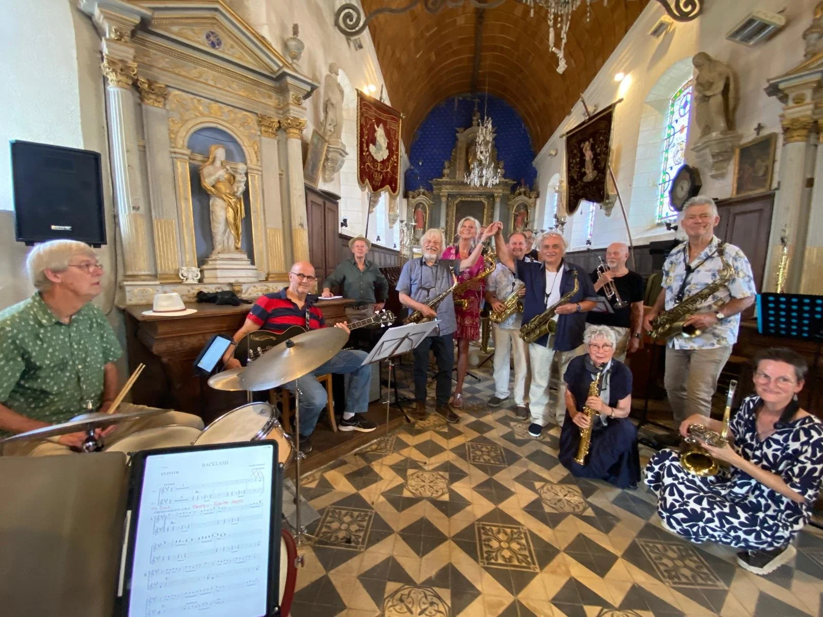 Een groep muzikanten poseert in een kerk, sommige met instrumenten zoals saxofonen, gitaren en drums, terwijl ze tijdens een oefening of optreden staan. Mensen lachen en lijken in een goede stemming.