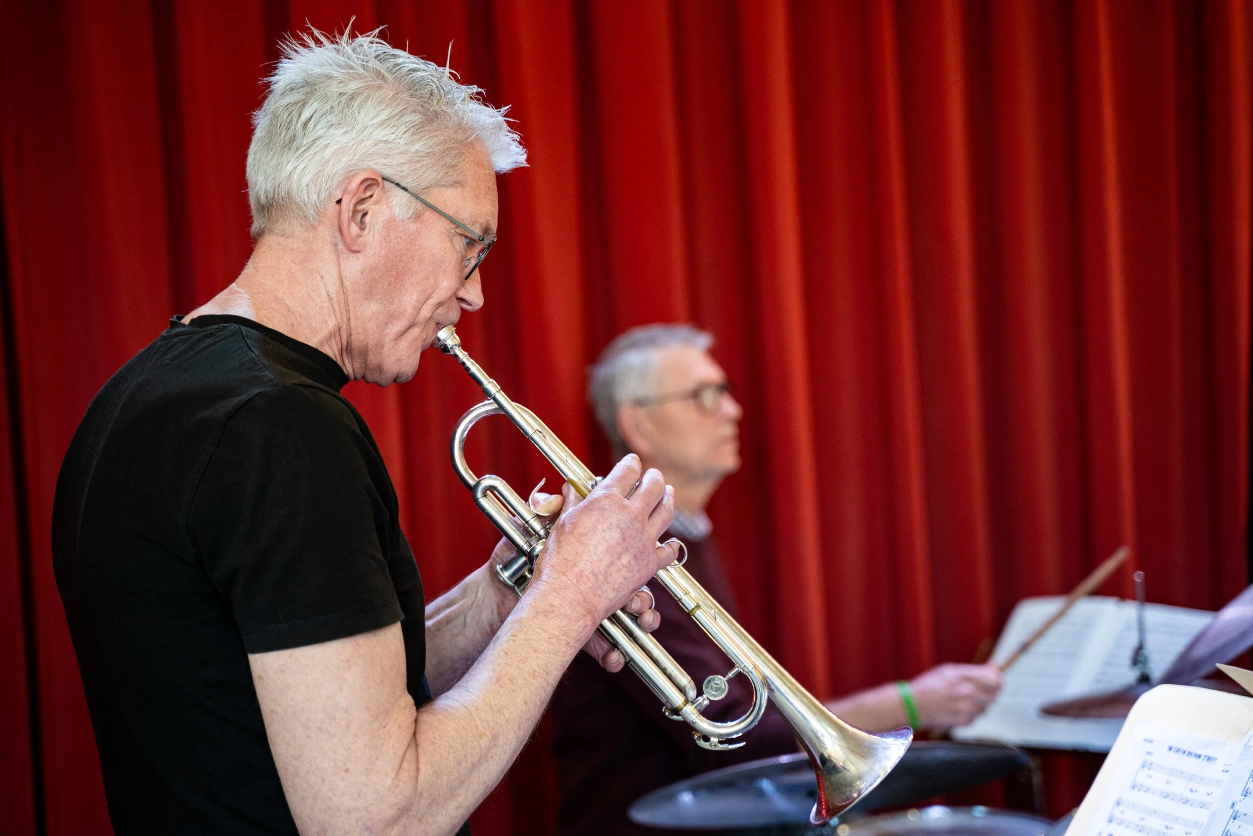 Man speelt de trompet, met een muzikale bladzijde voor zich, op de achtergrond een andere muzikant tegen een rood gordijn.